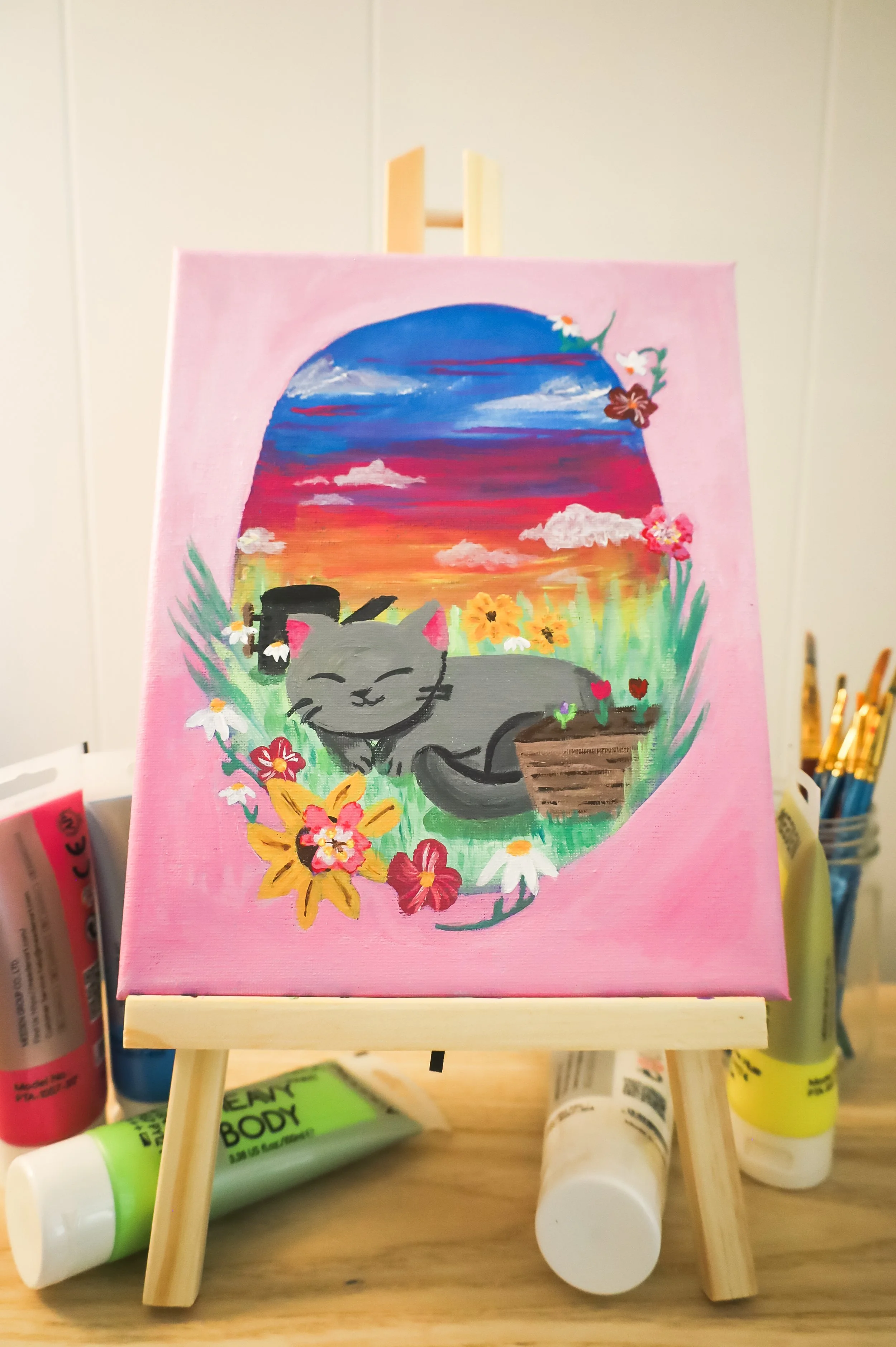 A miniature canvas painting on an easel depicts a gray cat with pink ears, relaxing among colorful flowers with a sunset sky in the background.