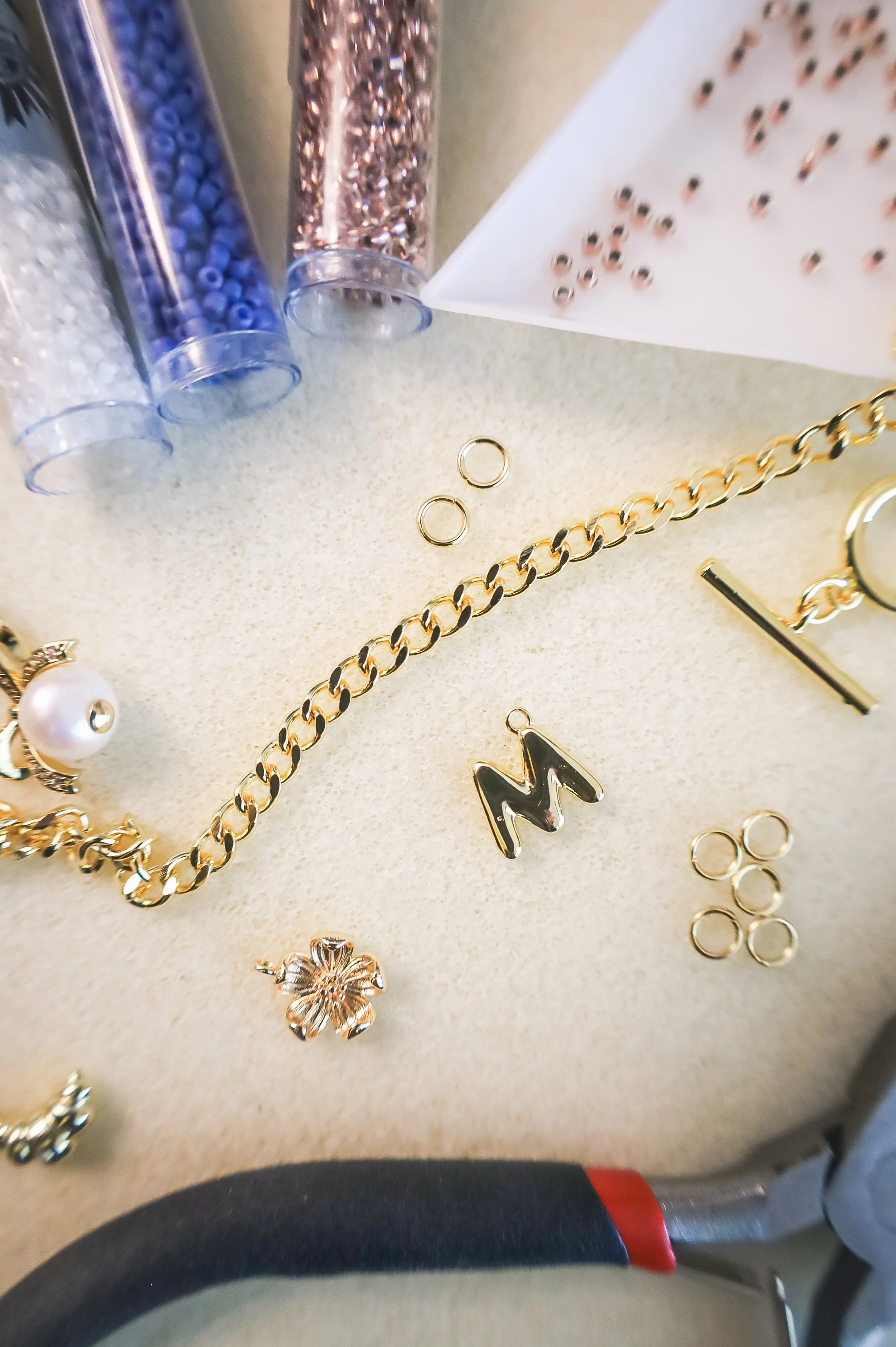 Jewelry-making supplies including a gold chain, letter 'M' pendant, flower charm, pearl earring, small gold rings, and small containers of colorful beads on a beige surface.