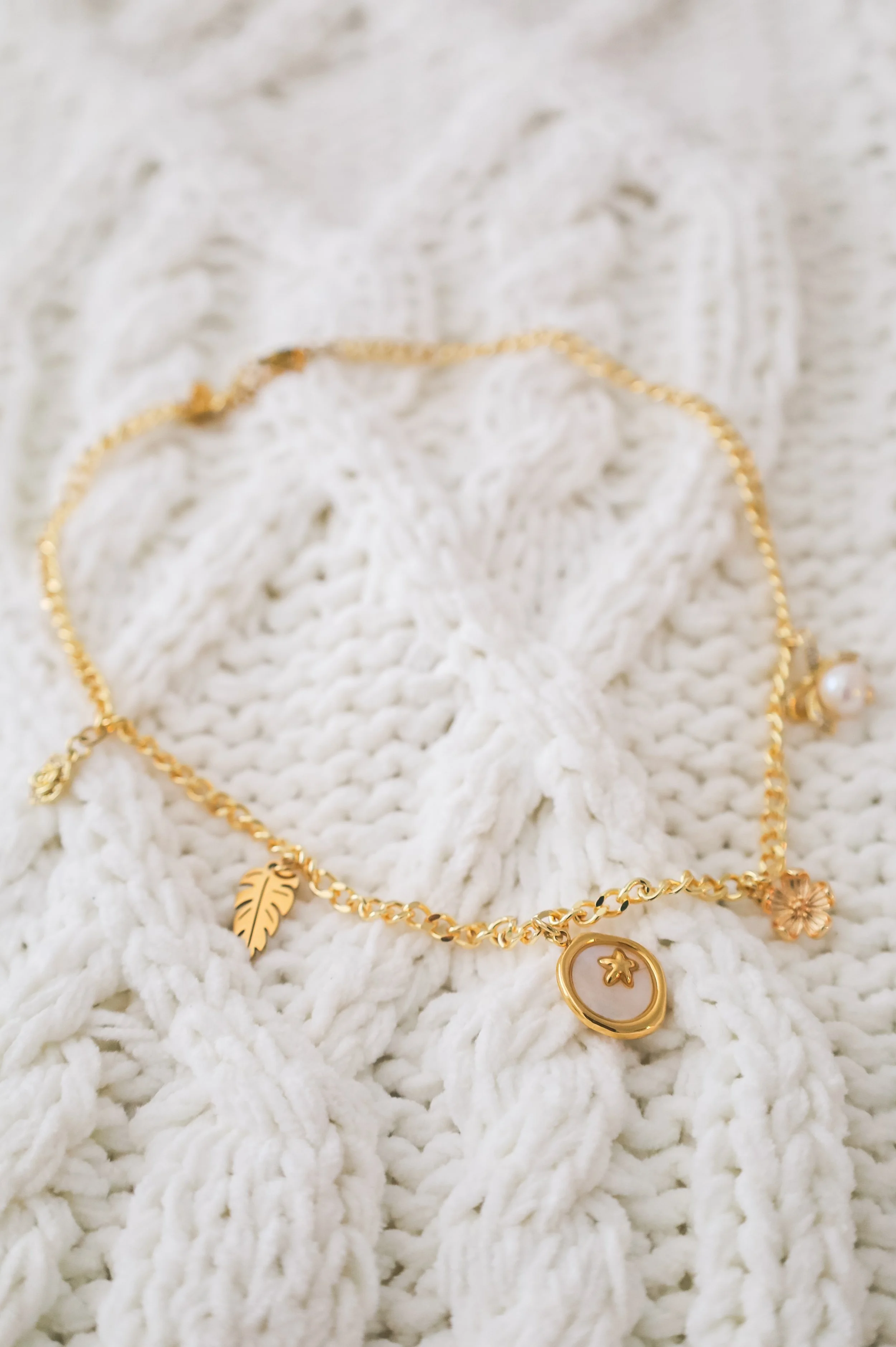 A gold chain bracelet with charms including a leaf, a star in a circle, and a flower, resting on a white knitted fabric.