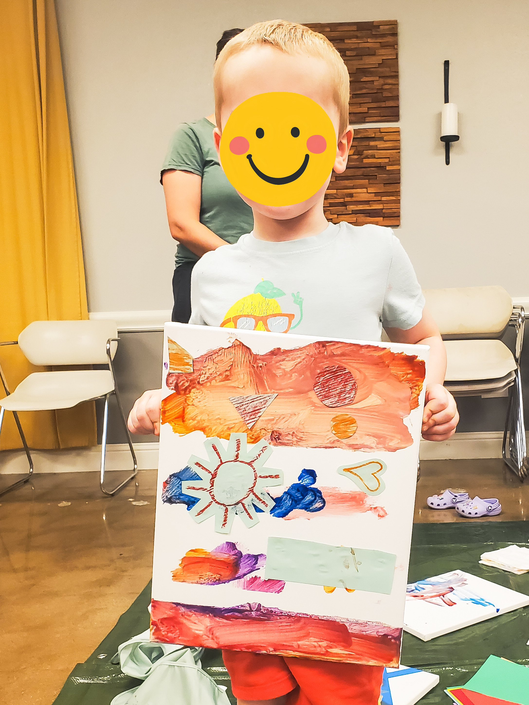 Child holding a colorful painted poster with drawings of a sun, a heart, and other shapes, while a person stands behind in a room with chairs, a wall with a wooden decorative piece, and yellow curtains.
