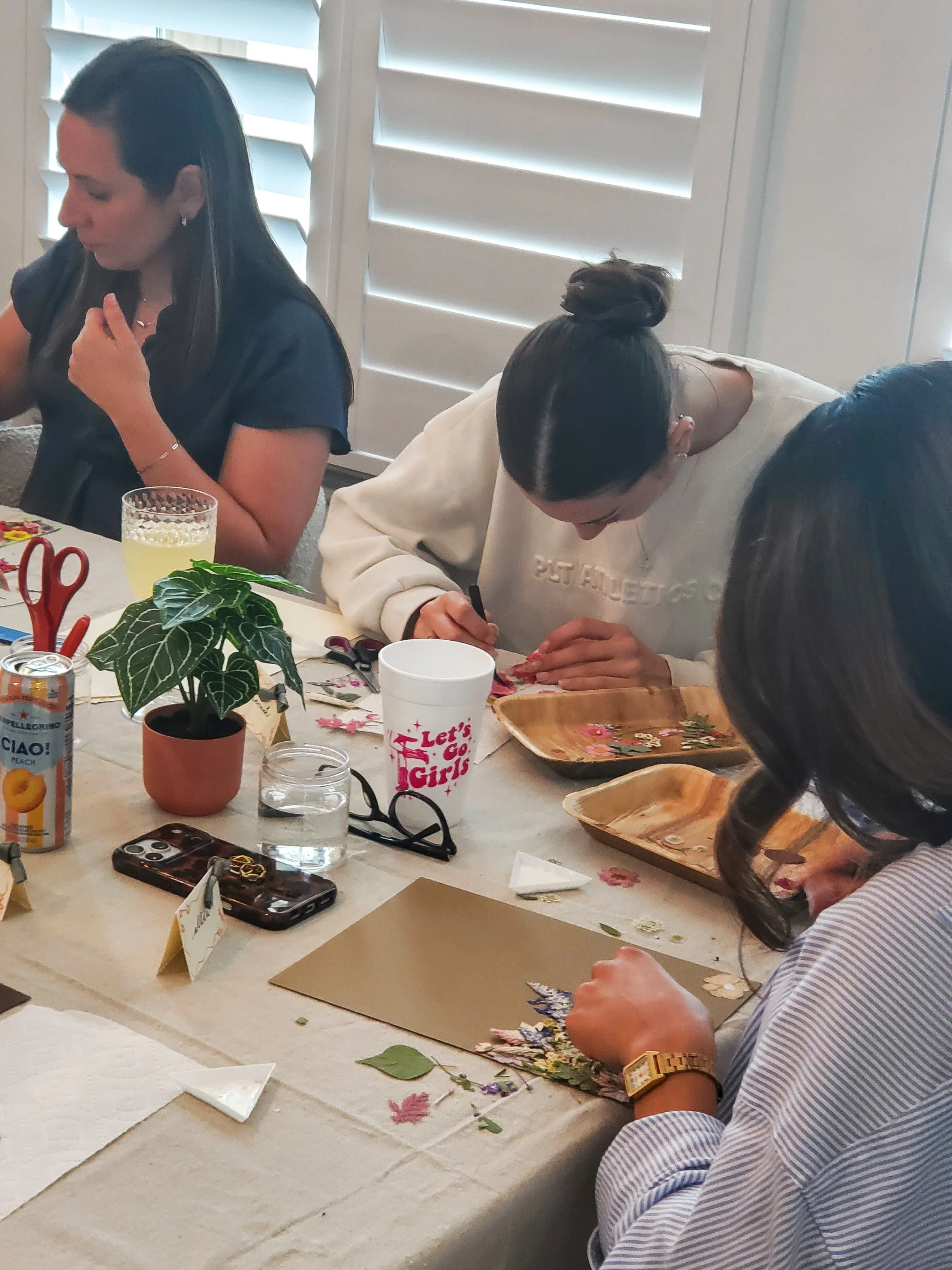 People sitting at a table engaged a pressed flower art project, with flowers and leaves, scissors, and crafting supplies.
