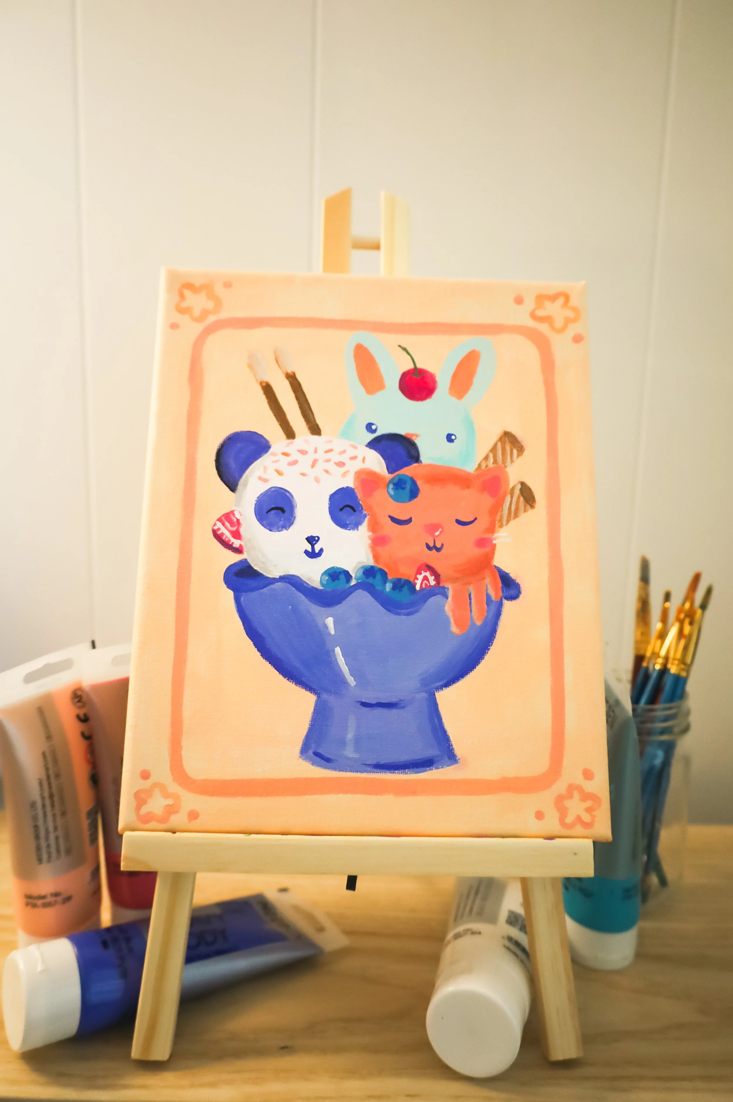Colorful illustration of a panda, bunny, and cat inside a blue bowl with cookies and a cherry on top, displayed on a small easel.