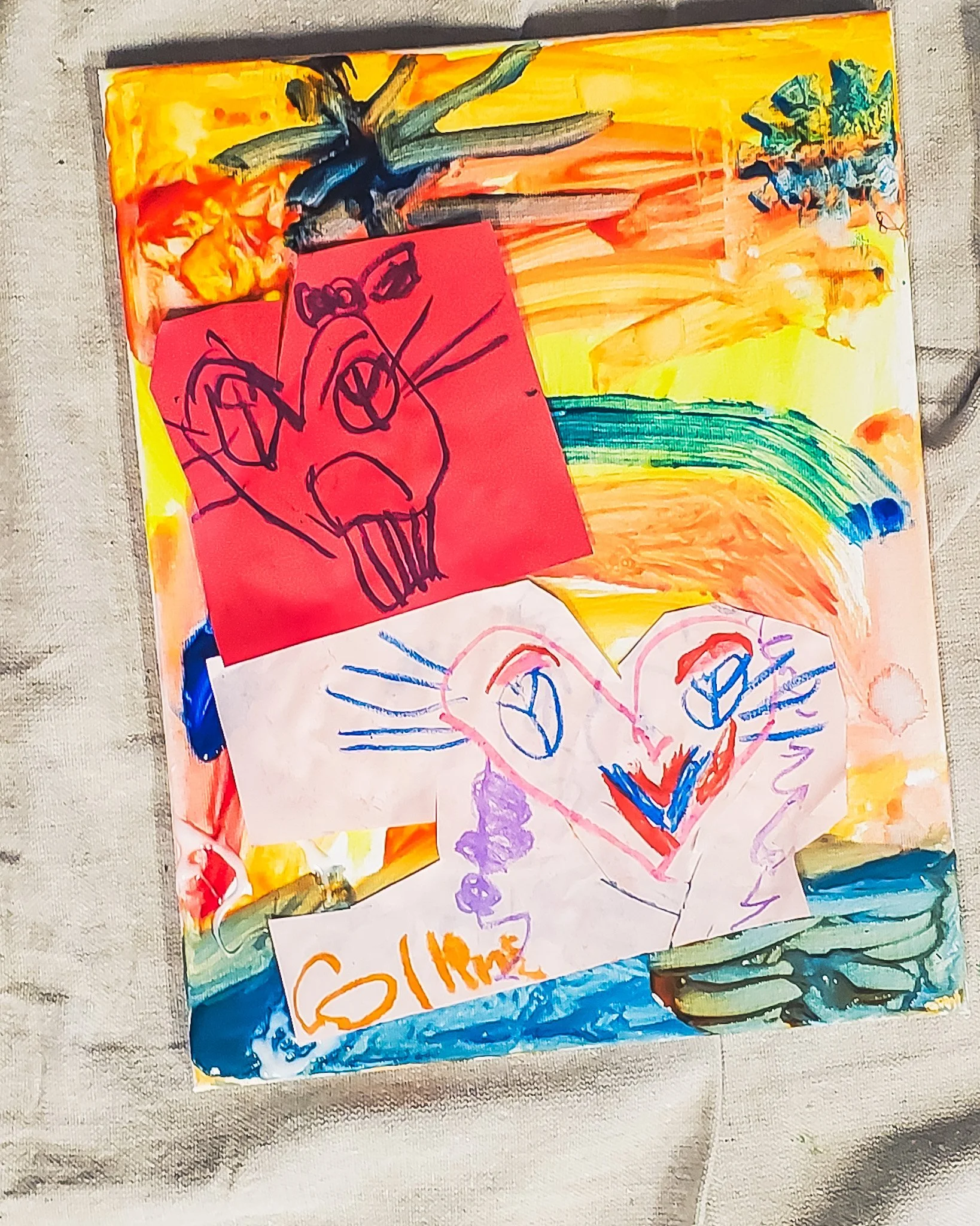 A colorful children's art display on a fabric surface, featuring various drawings and paintings including a rainbow, a red paper with a child's drawing, and a white piece of paper with abstract marks.
