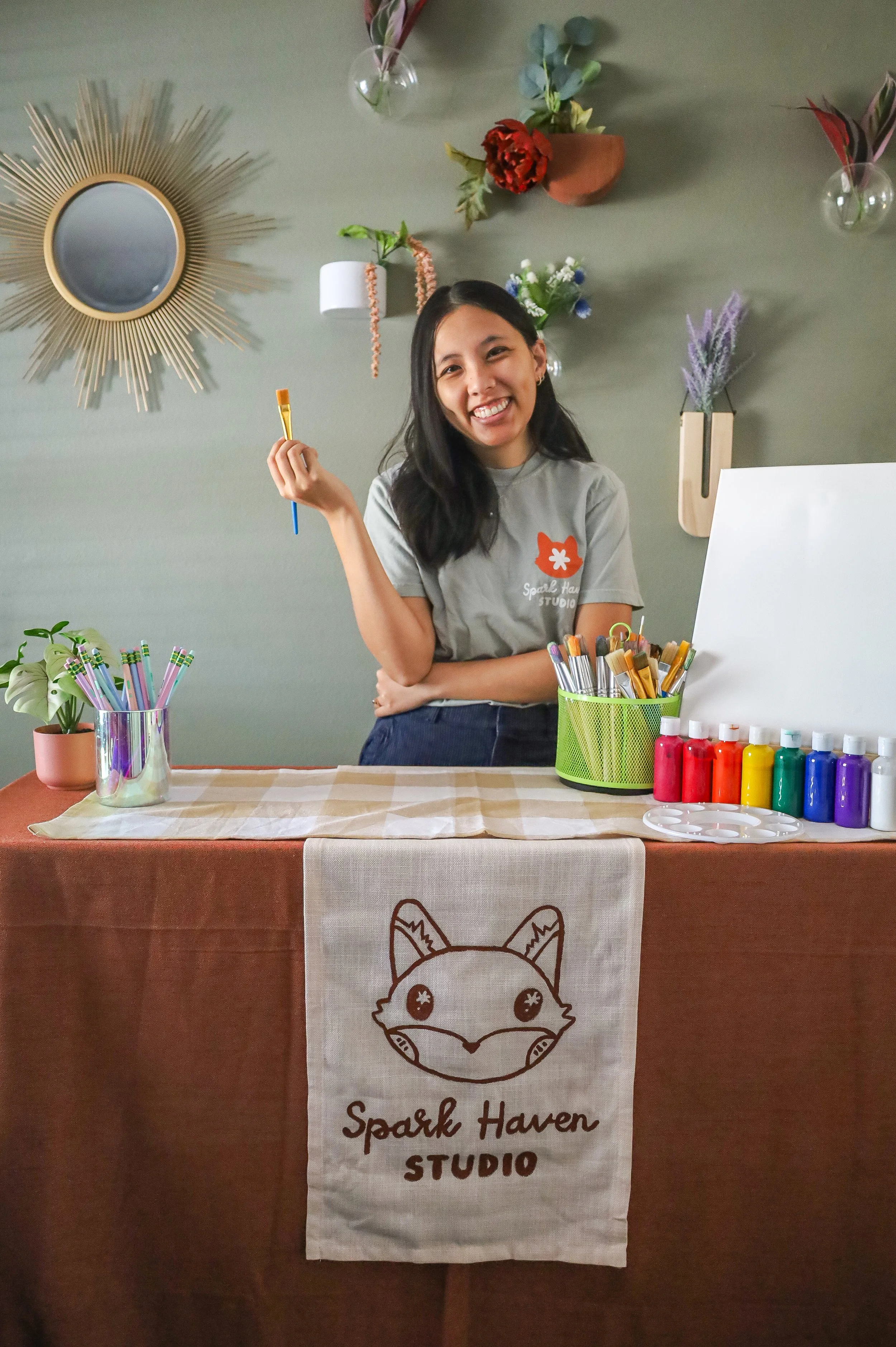 A smiling woman standing behind a table with art supplies, including paintbrushes and bottles of paint, in a decorated room with wall-mounted vases and a round mirror, promoting Spark Haven Studio.