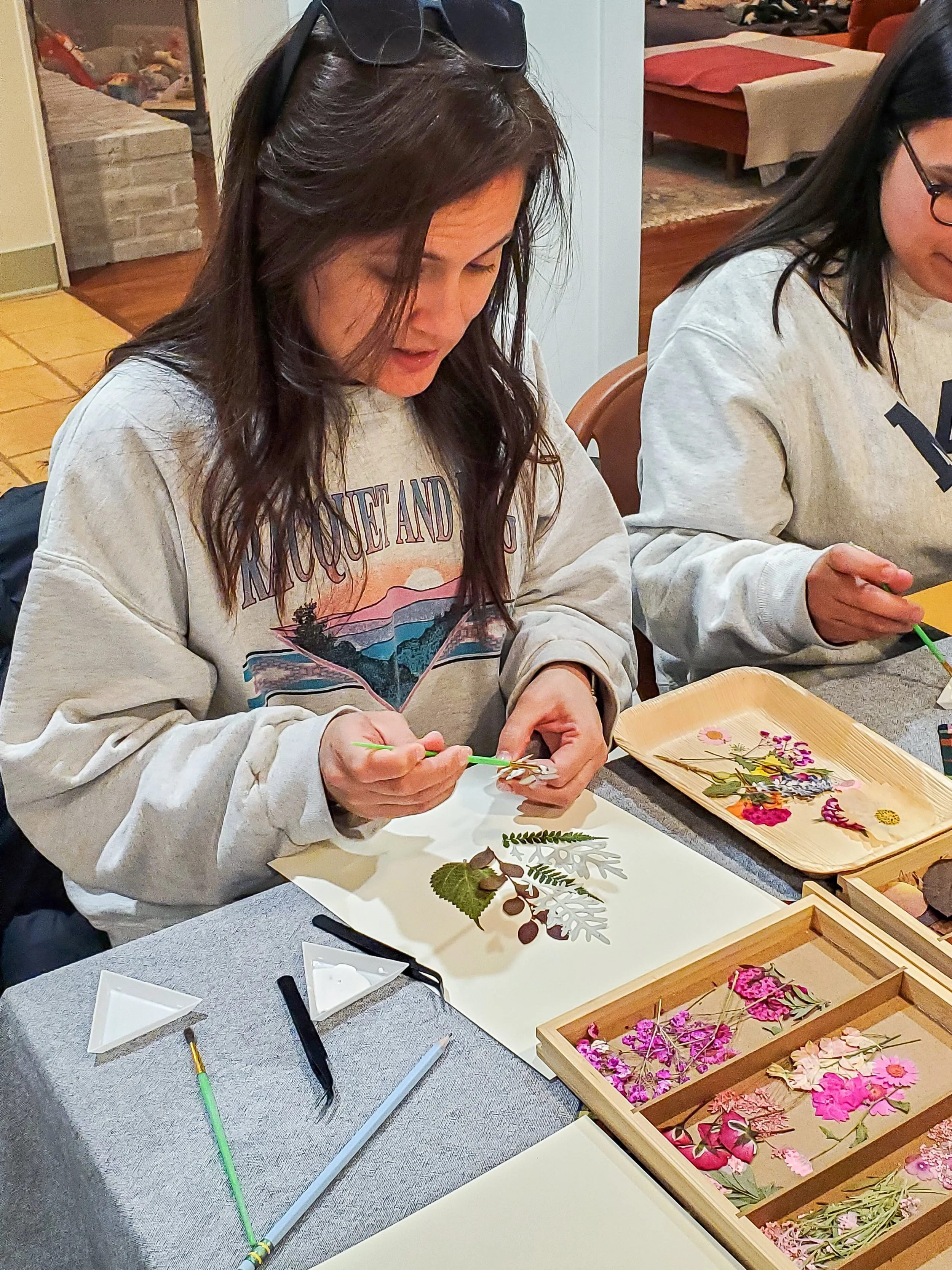 Woman creating pressed flower art at a craft table, with various flowers and leaves laid out.