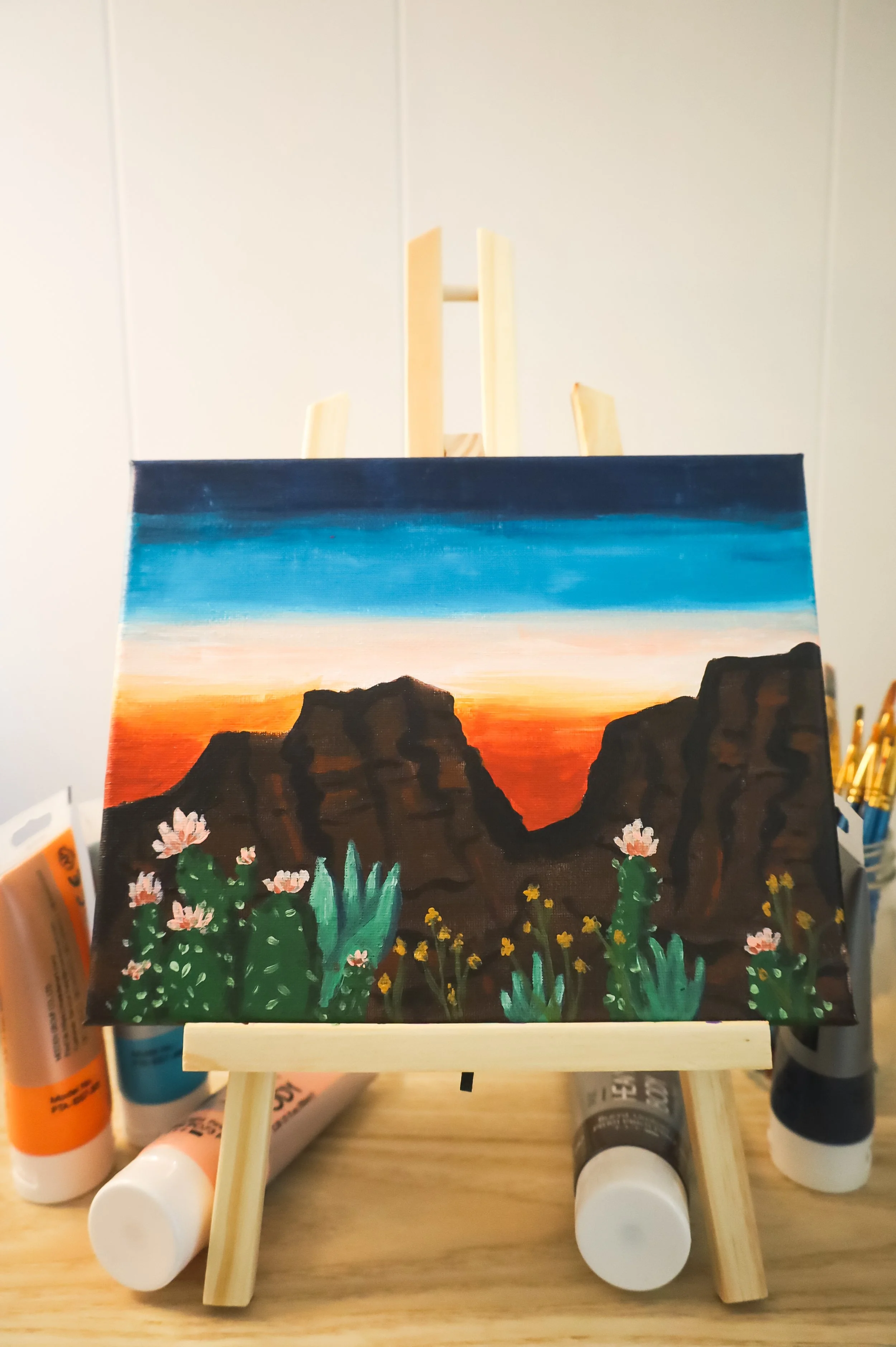 A small painting of a desert landscape with cacti, rocks, and a sunset sky, on a wooden easel.