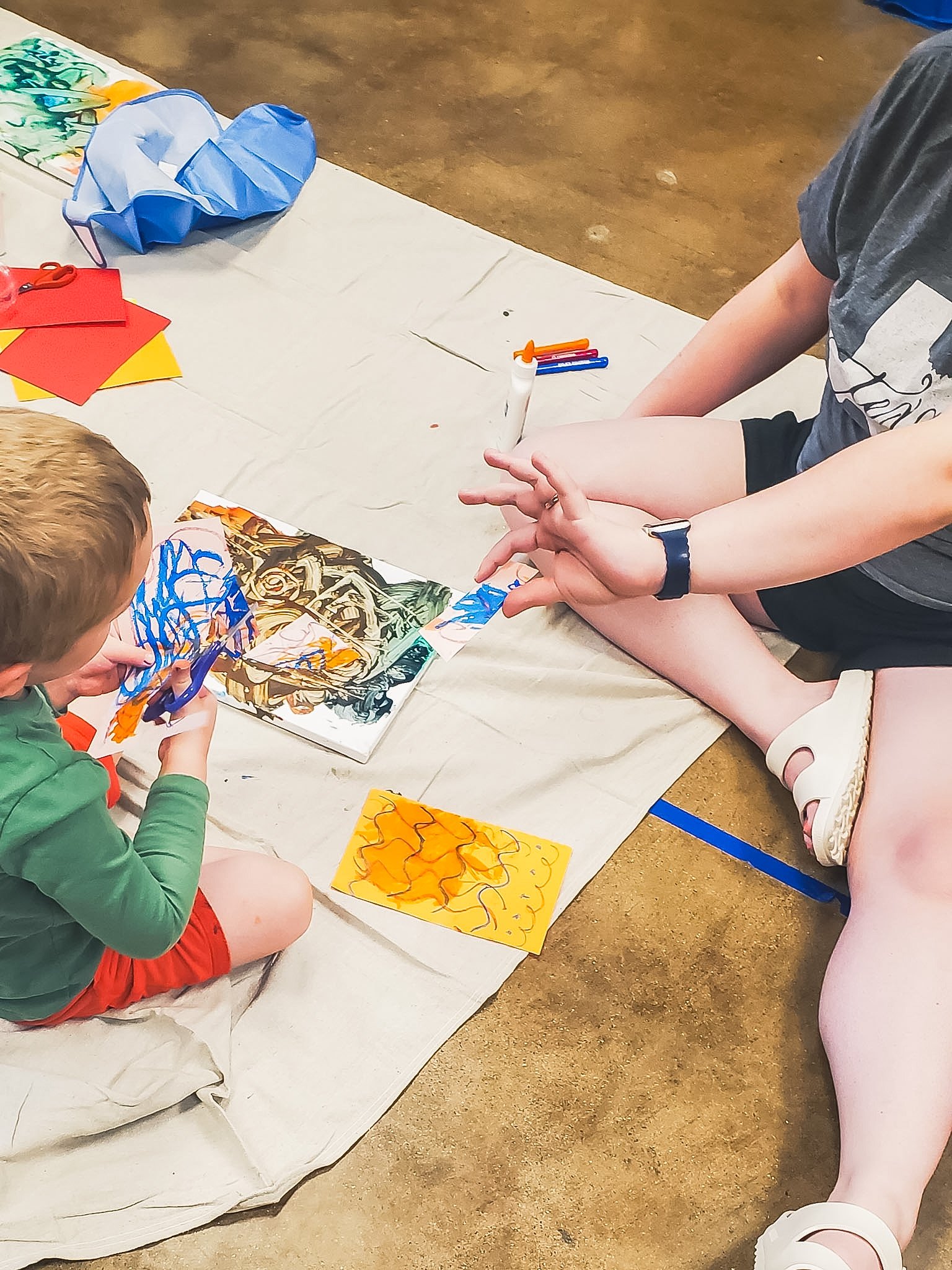 A child sitting on a cloth-covered floor creating artwork with paint, canvas, paper, scissors, and glue. The cloth-covered floor has colorful papers and art supplies.