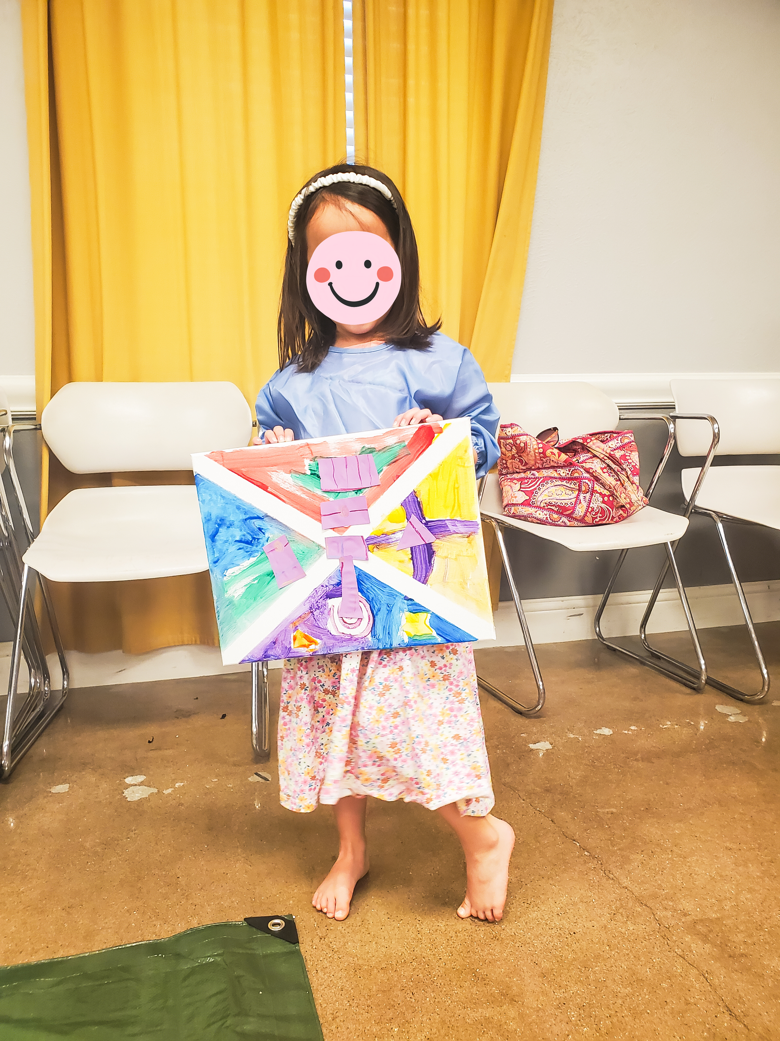 A young girl stands on a tan floor, holding a colorful painted canvas with various colors and shapes. She wears a floral long skirt, a light blue smock, and has a headband. There are chairs, a yellow curtain, and a bag on a chair in the background.