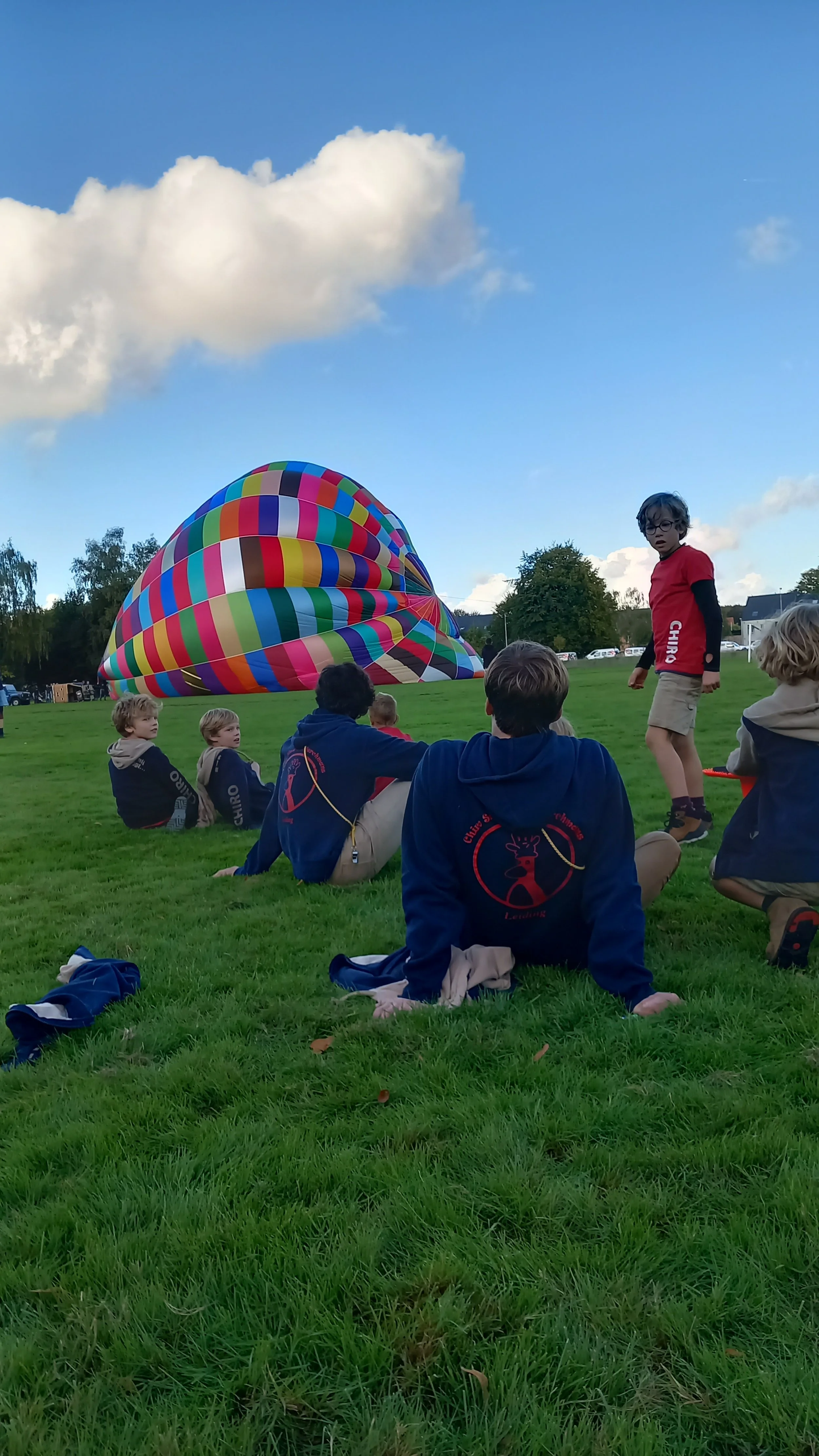 Children sitting on grass field, some wearing matching shirts with a deer logo, observing a colorful hot air balloon lying on the ground against a blue sky with clouds.