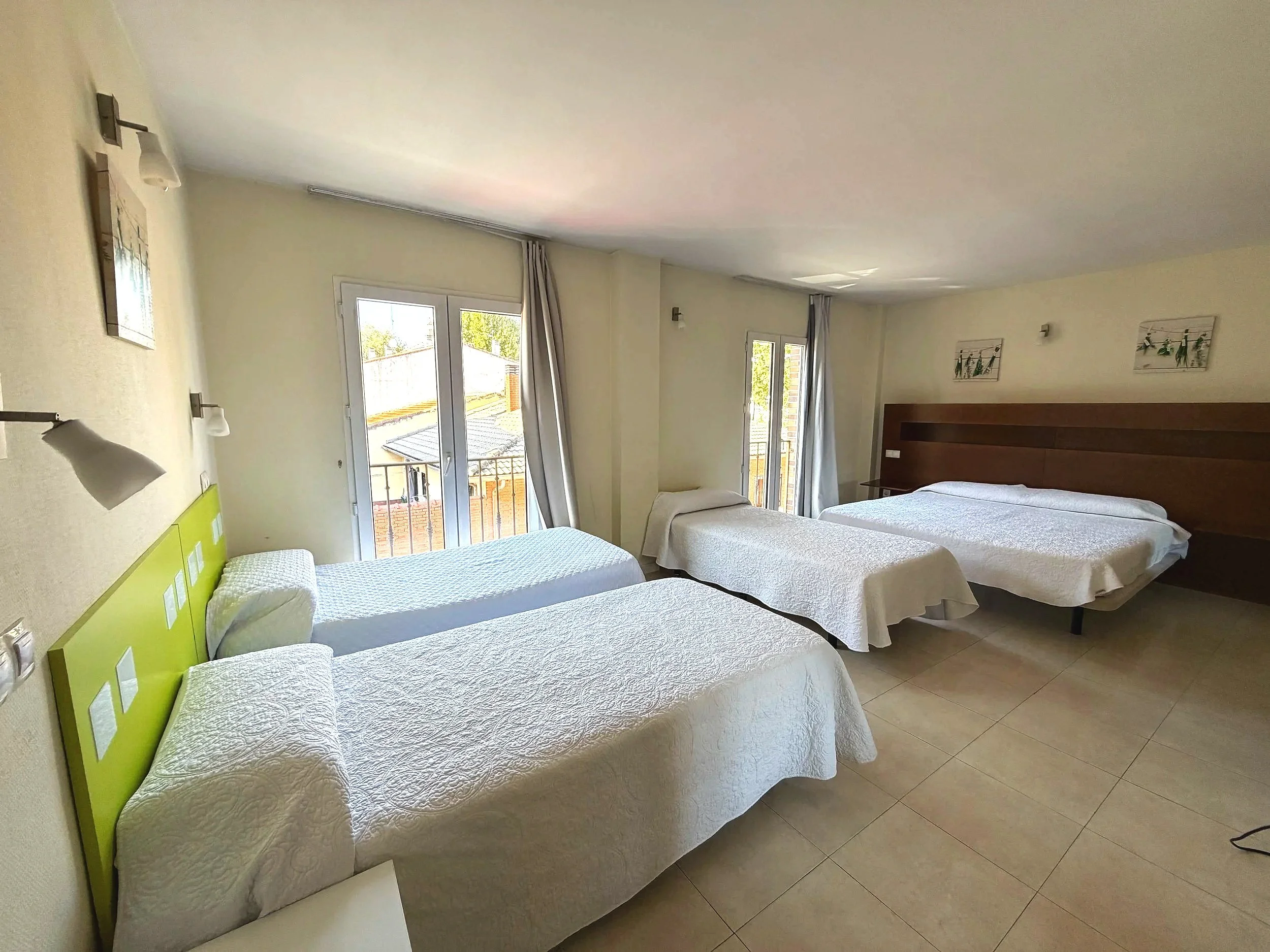 Habitación con cuatro camas de matrimonio con colchas blancas, paredes de color crema, ventanas con cortinas grises y arte decorativo en las paredes.