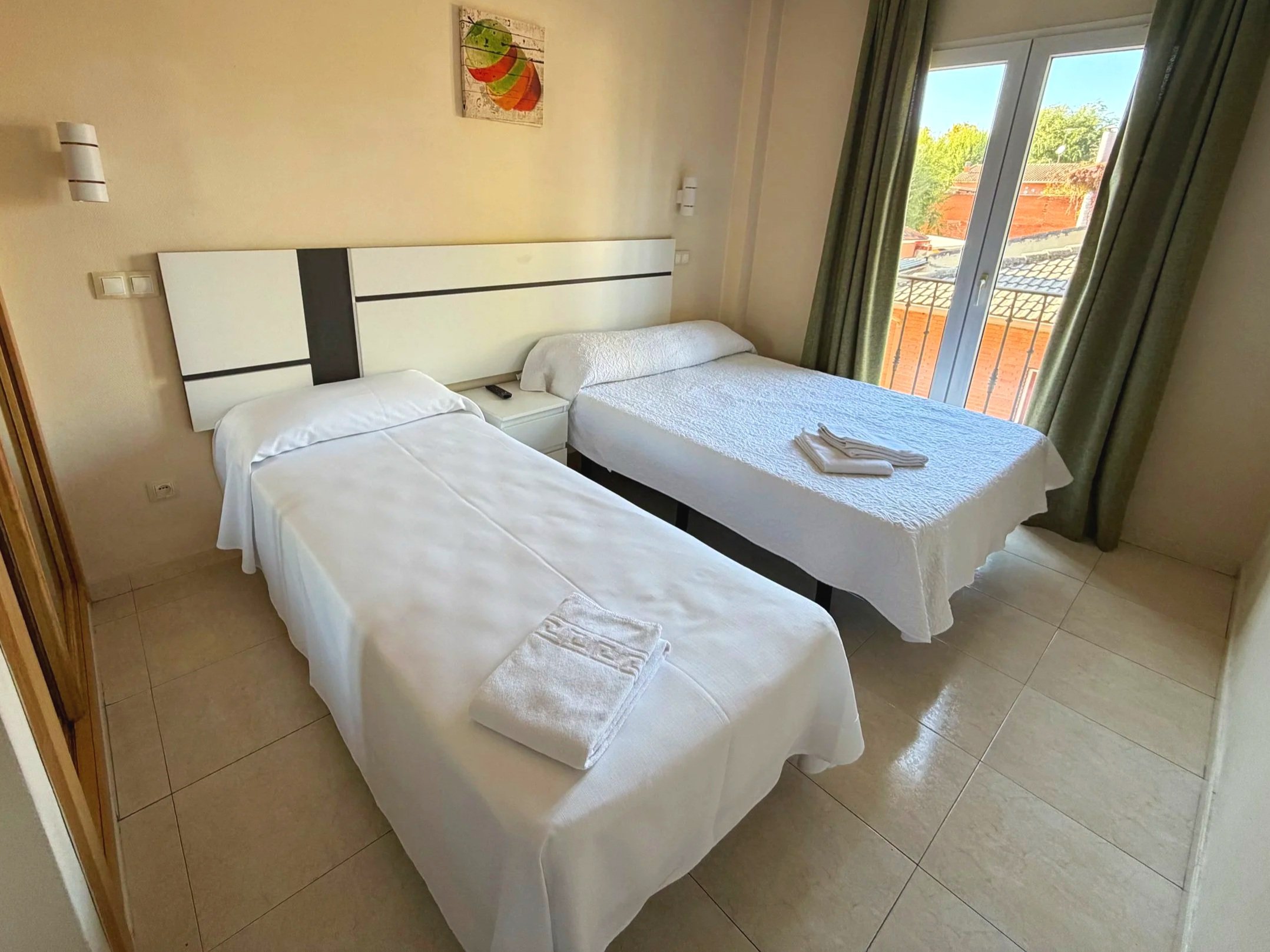 Habitación de hotel con dos camas, una con sábanas blancas, y la otra con mantas blancas y toallas en la cama, ventana con cortinas verdes, y cuadros decorativos en la pared.