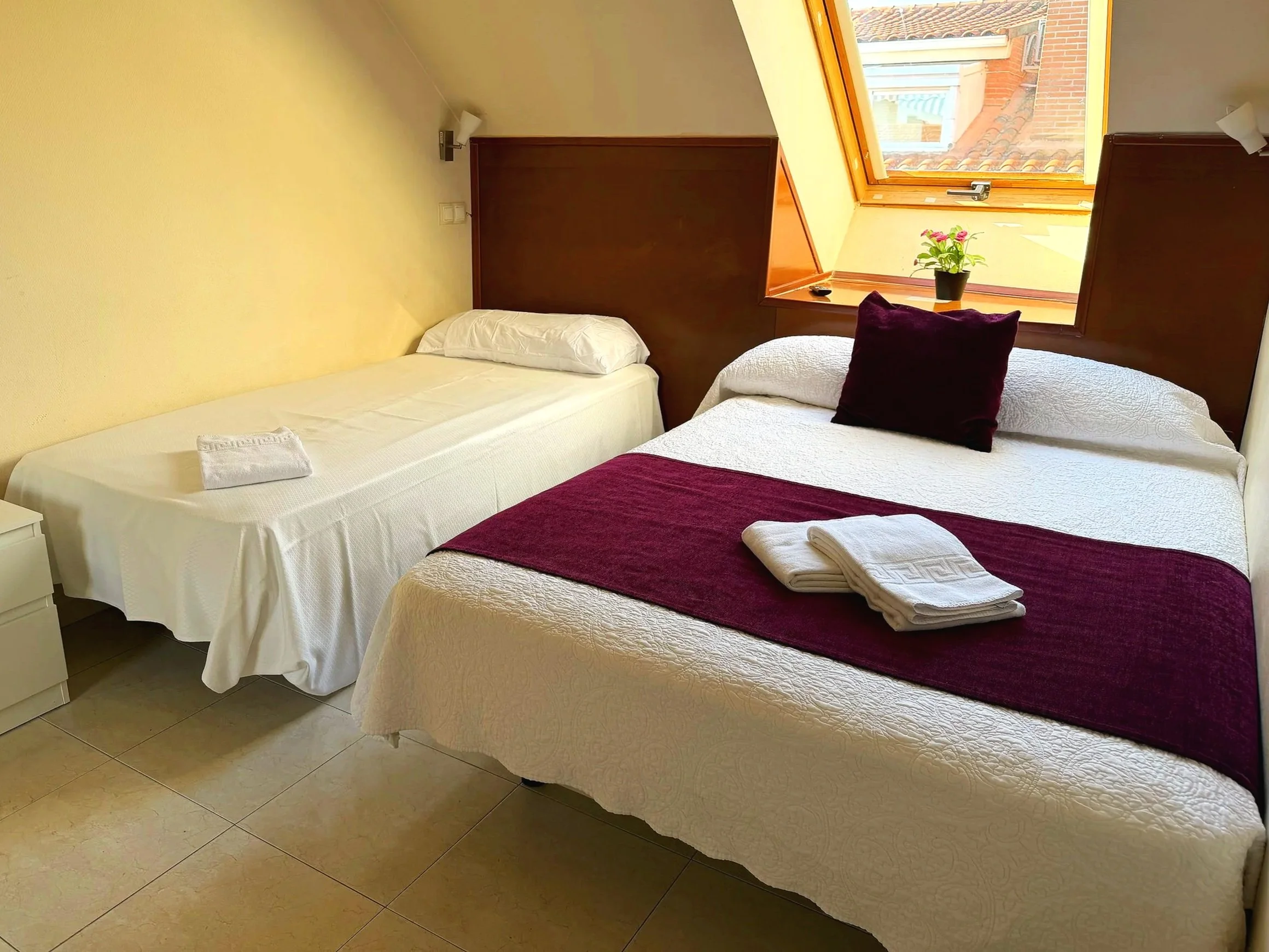 Habitación con dos camas, una doble y una individual, cerca de una ventana con vista a tejados y cielo, decorada con mantas y toallas blancas, una planta en la ventana y paredes de color crema.