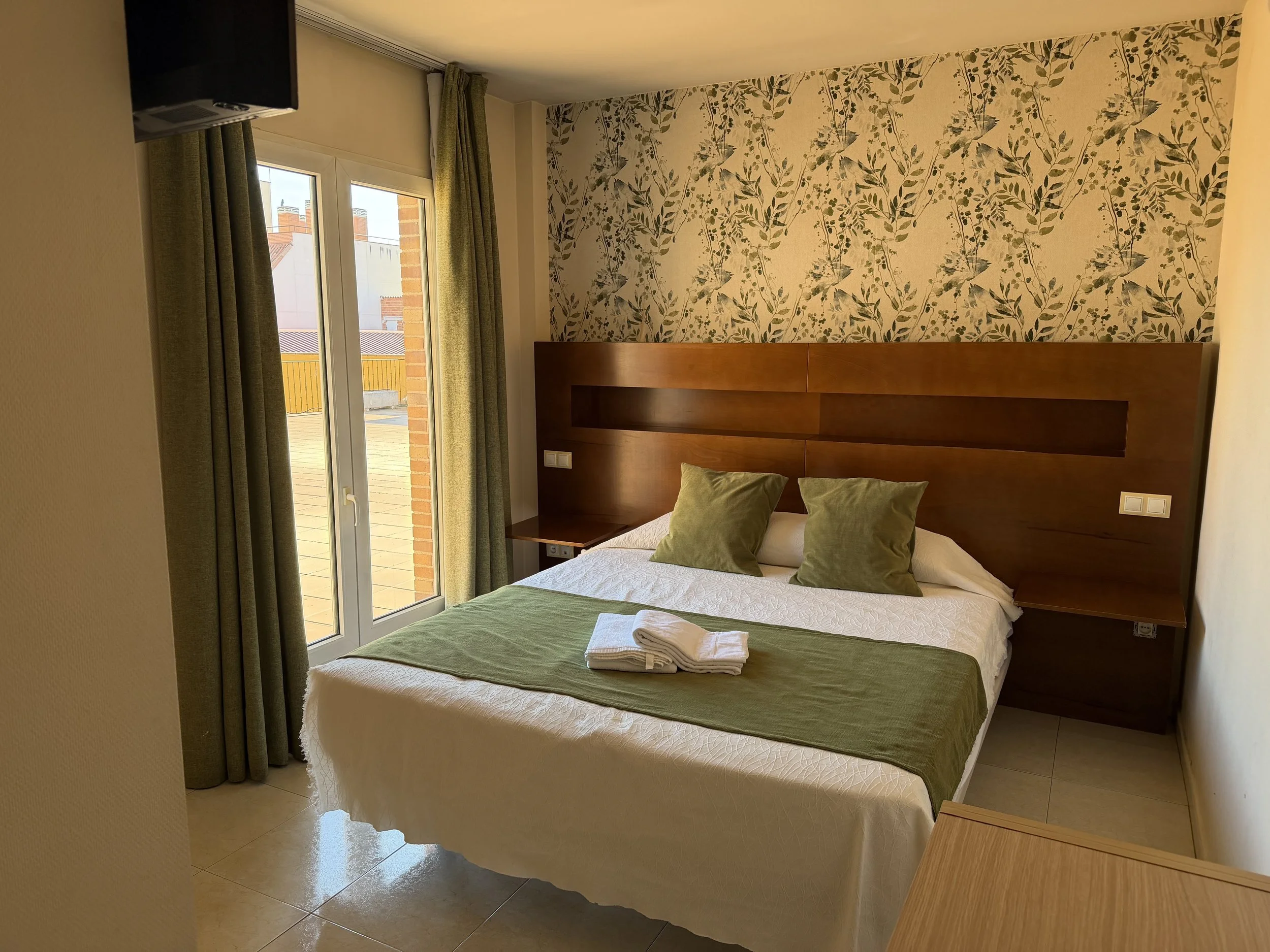 Habitación de hotel con cama doble, cabecera de madera y paredes decoradas con papel tapiz de hojas verdes, ventana con cortinas verdes y balcón con vistas a edificios.