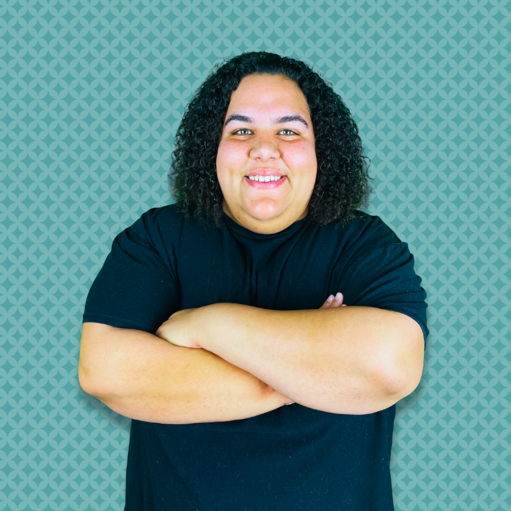 Person with curly hair wearing a black shirt standing with arms crossed in front of a patterned teal background.