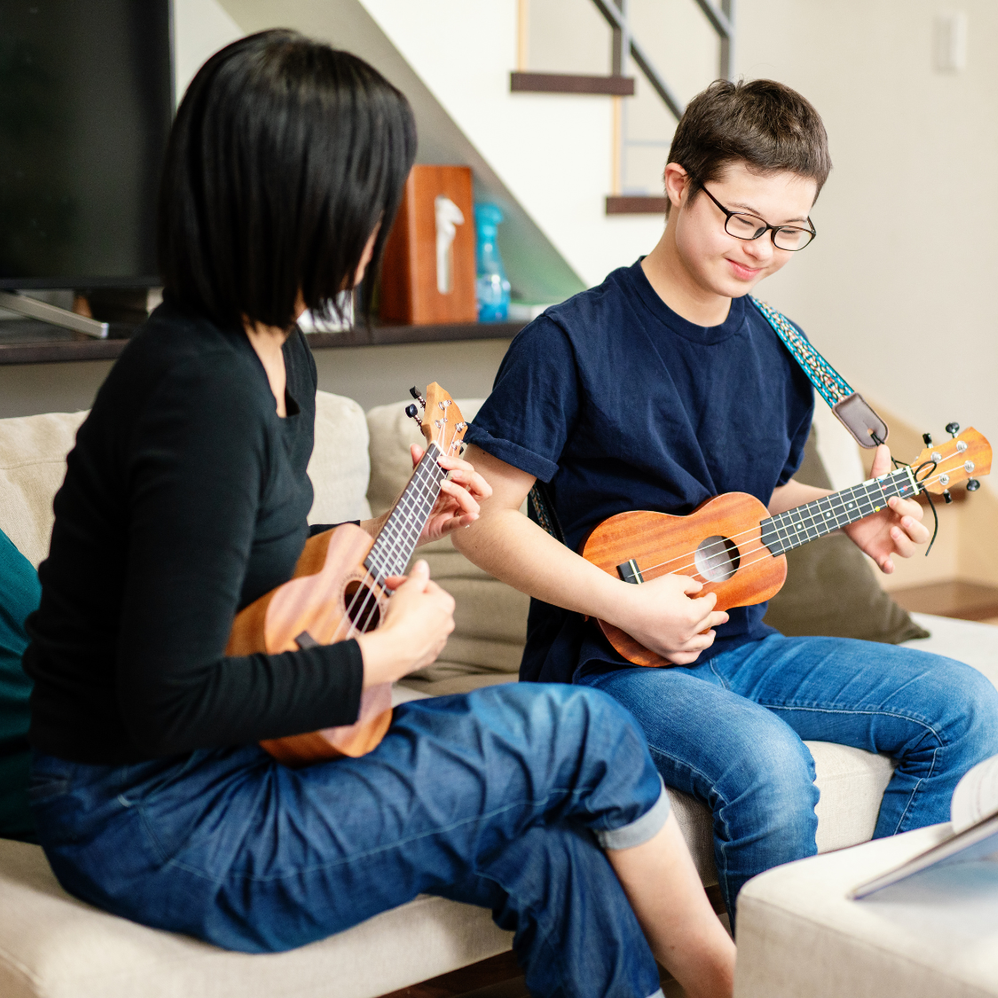 Two young people sitting on a couch playing ukuleles, smiling, in a cozy living room.