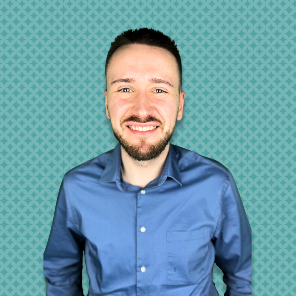 A man with short dark hair, a beard, and mustache, smiling in a blue button-up shirt, against a teal patterned background.