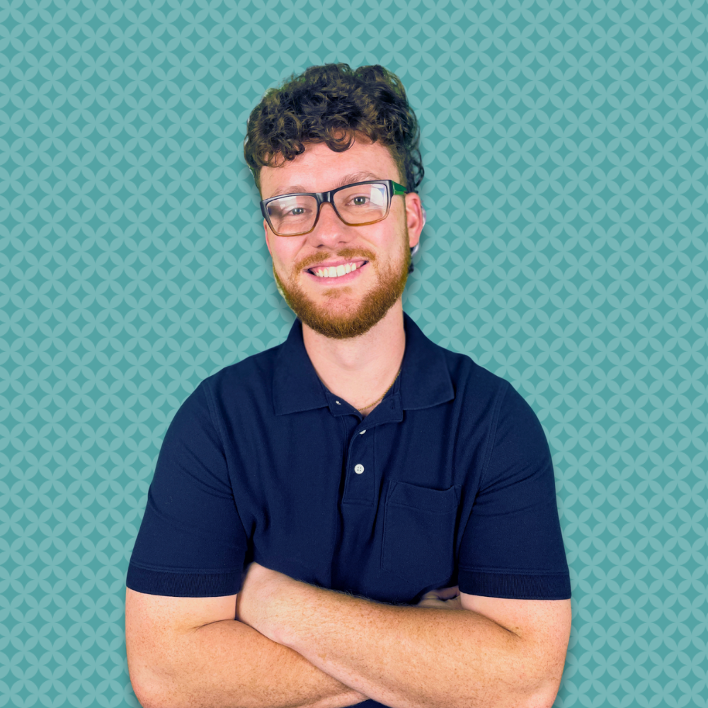 A man with curly hair, glasses, and a beard, smiling with arms crossed, wearing a navy blue polo shirt, standing in front of a teal patterned background.