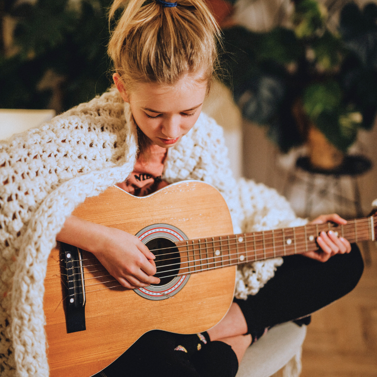A woman sitting indoors playing an acoustic guitar, wrapped in a chunky knit blanket