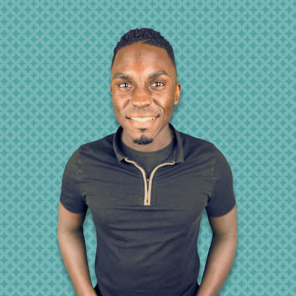 A smiling man with short curly hair, wearing a black zip-up shirt with a collar, standing in front of a turquoise patterned background.