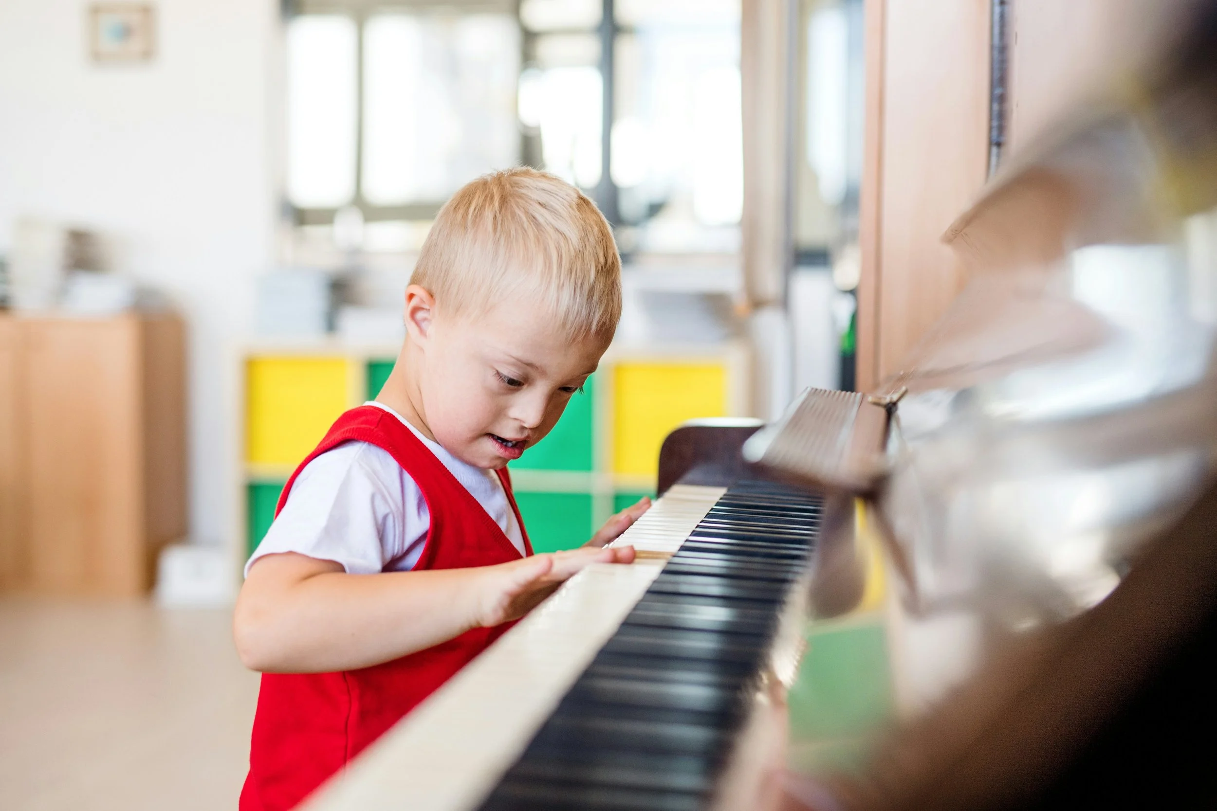 A young boy in a red vest playing a piano in a brightly lit classroom.