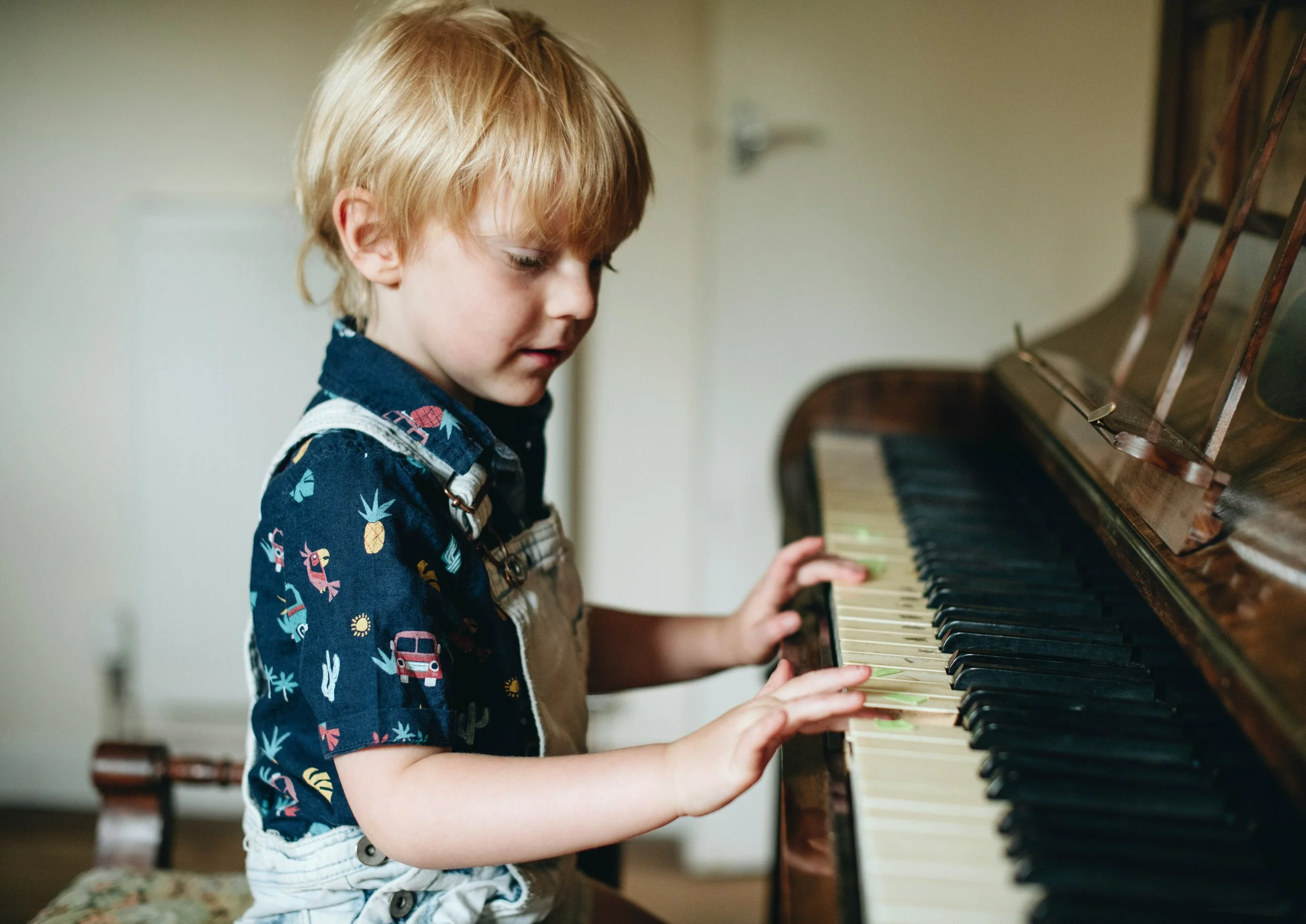 A young boy with blond hair playing the piano, wearing a navy shirt with colorful animal and vehicle print and beige overalls, sitting at a wooden upright piano.