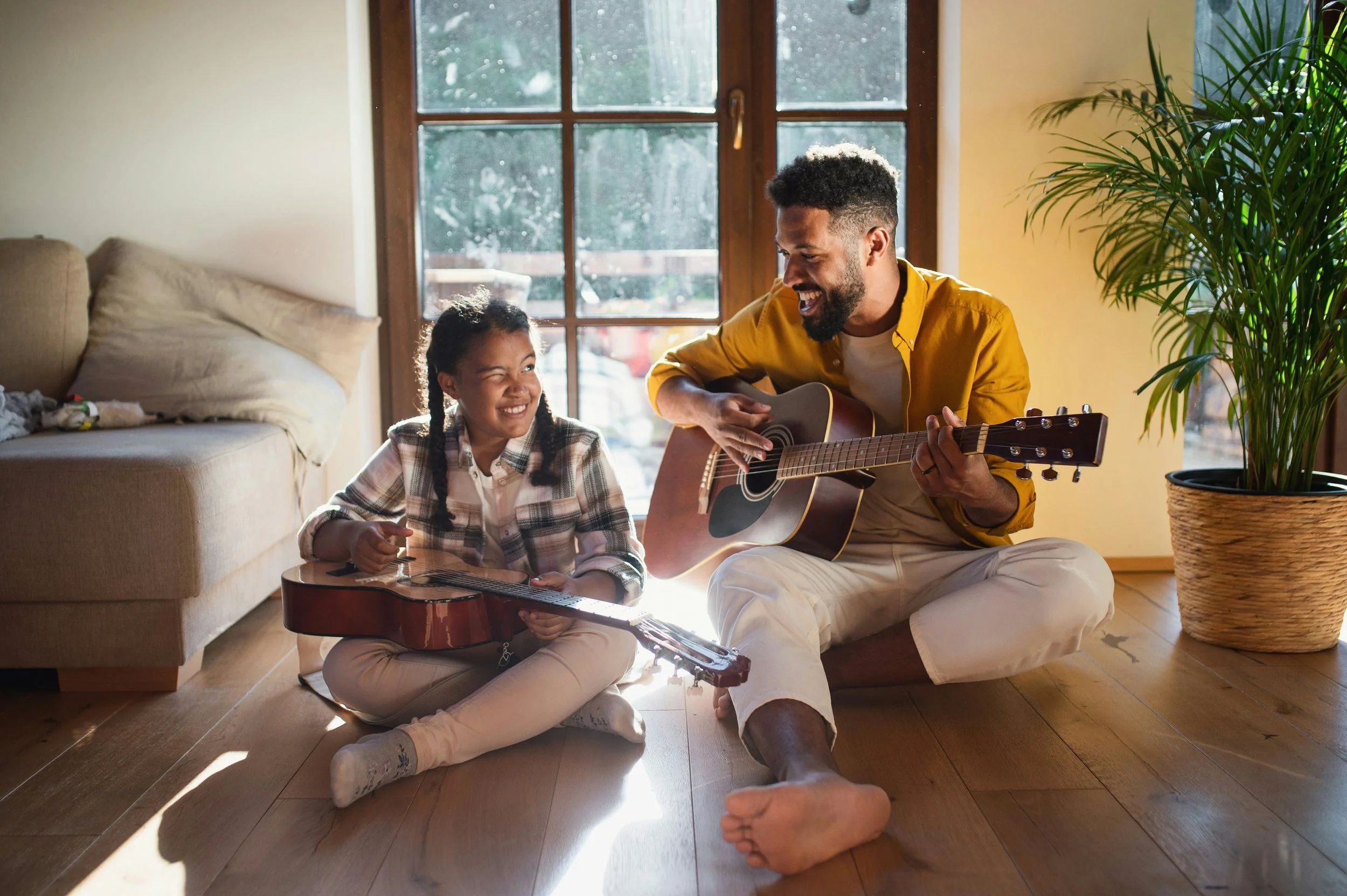 A man and a young girl sitting on the wooden floor in a sunlit room, playing guitars and smiling at each other.