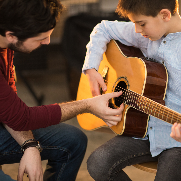 A young boy learning to play an acoustic guitar with an instructor in a music classroom.