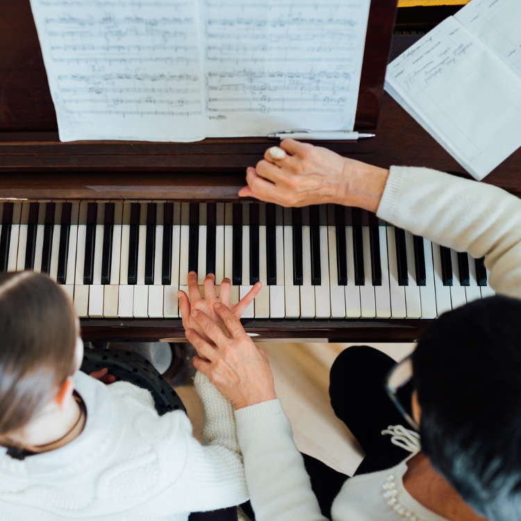 A child and an adult sitting together at a piano, with the child's hand on the keys and the adult guiding them, sheet music on the piano stand.