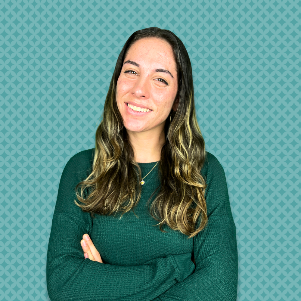 A young woman with long wavy hair wearing a dark green sweater, smiling with arms crossed, standing against a teal patterned background.