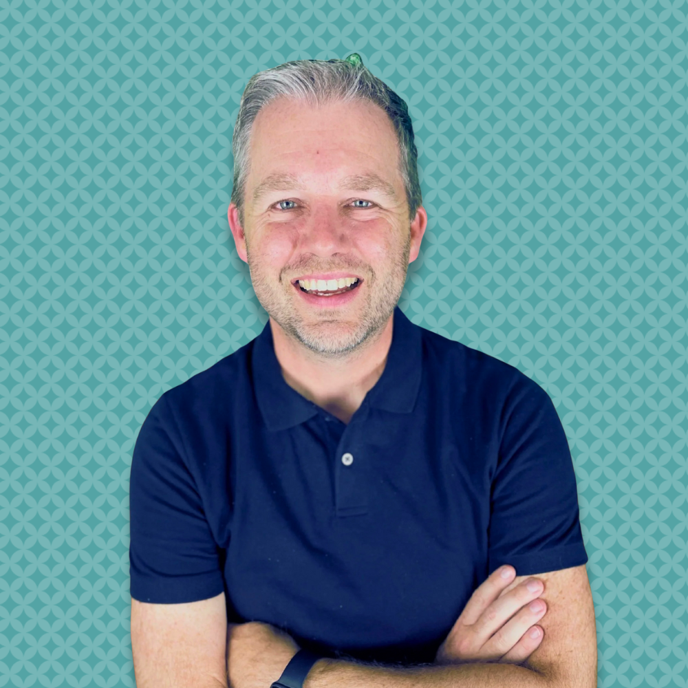 A smiling man with short gray hair and a beard, wearing a navy blue polo shirt, standing with arms crossed against a teal patterned background.