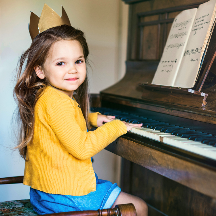 A young girl with long hair, wearing a yellow sweater and a paper crown, playing the piano and looking at the camera.