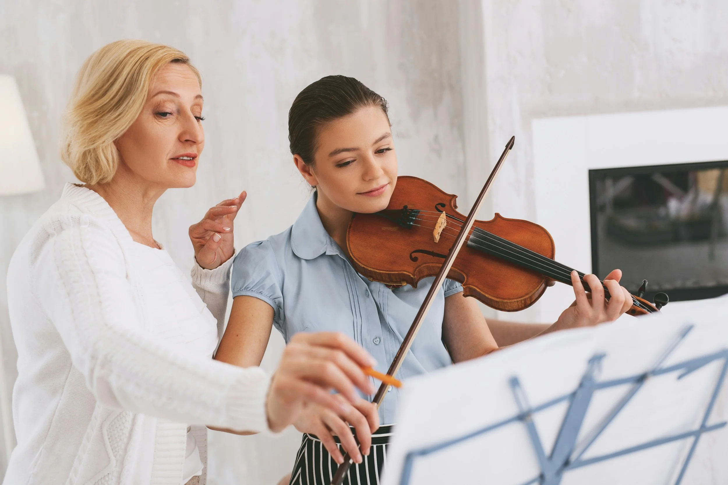 An older woman instructs a young girl playing the violin in a music lesson.
