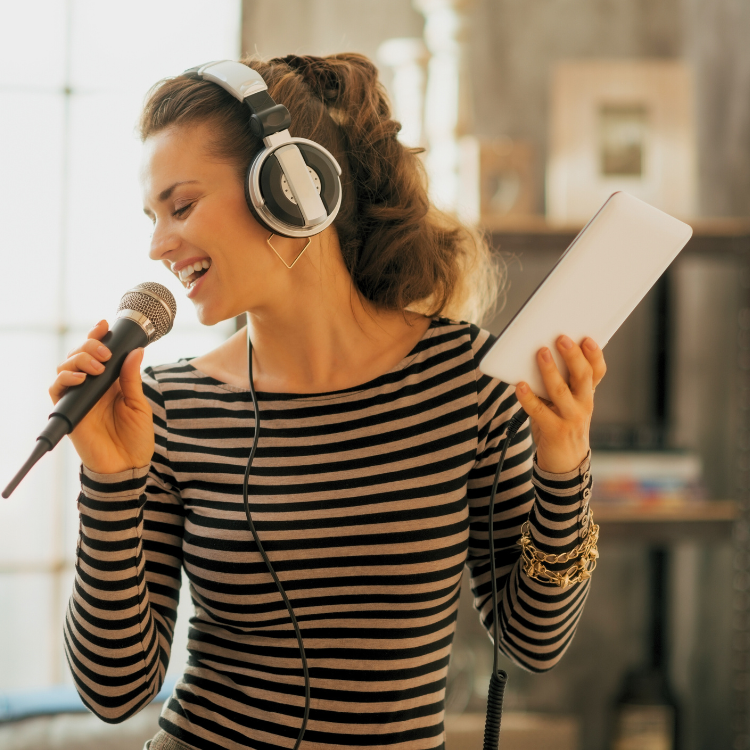 A woman with curly hair wearing headphones and a striped shirt singing into a microphone while holding a tablet in her other hand.
