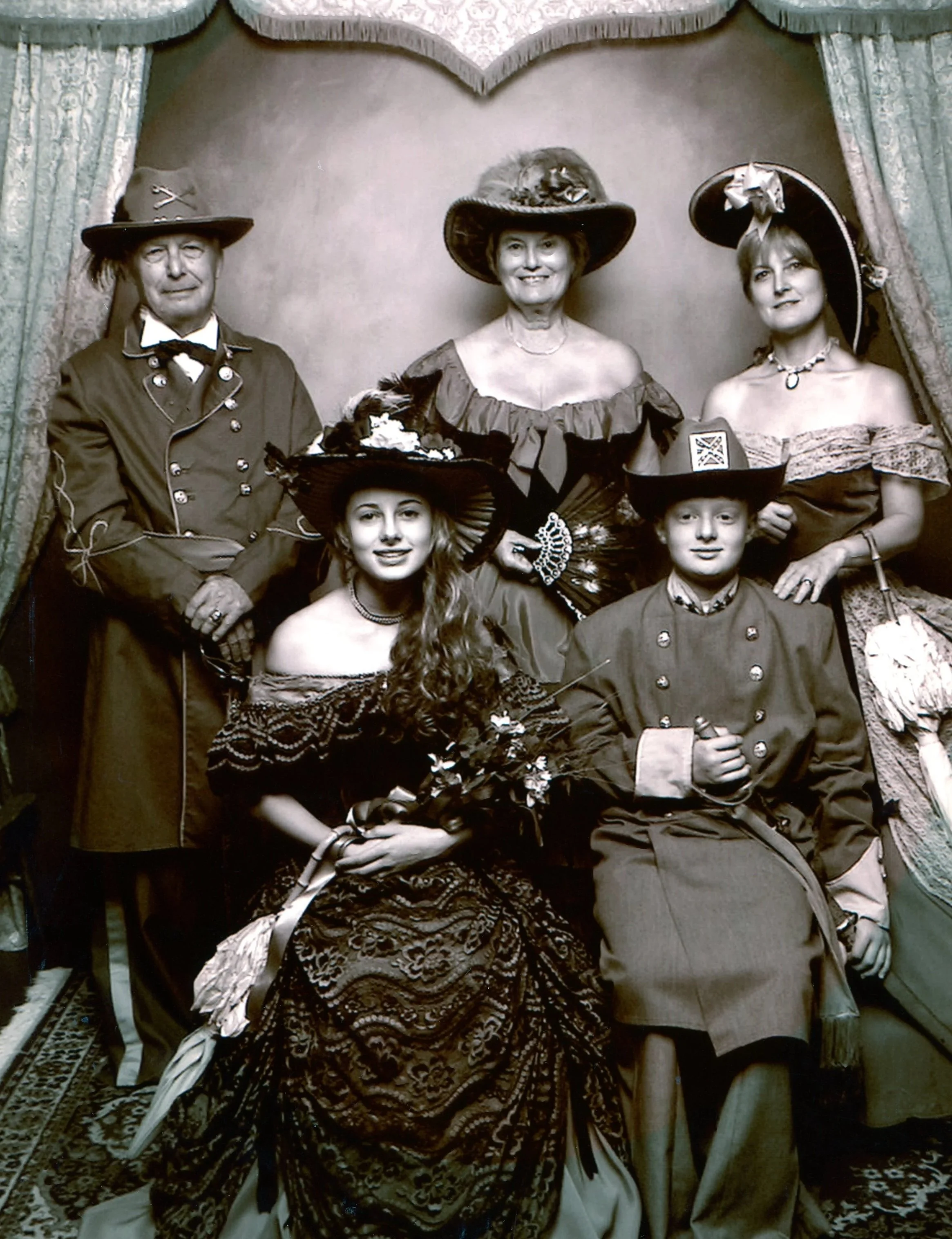 A vintage sepia photograph of six people dressed in Victorian-style costumes, with three standing in the back row and two seated in the front. They are all wearing elaborate hats and period-appropriate clothing, and are posed in front of a decorative backdrop.