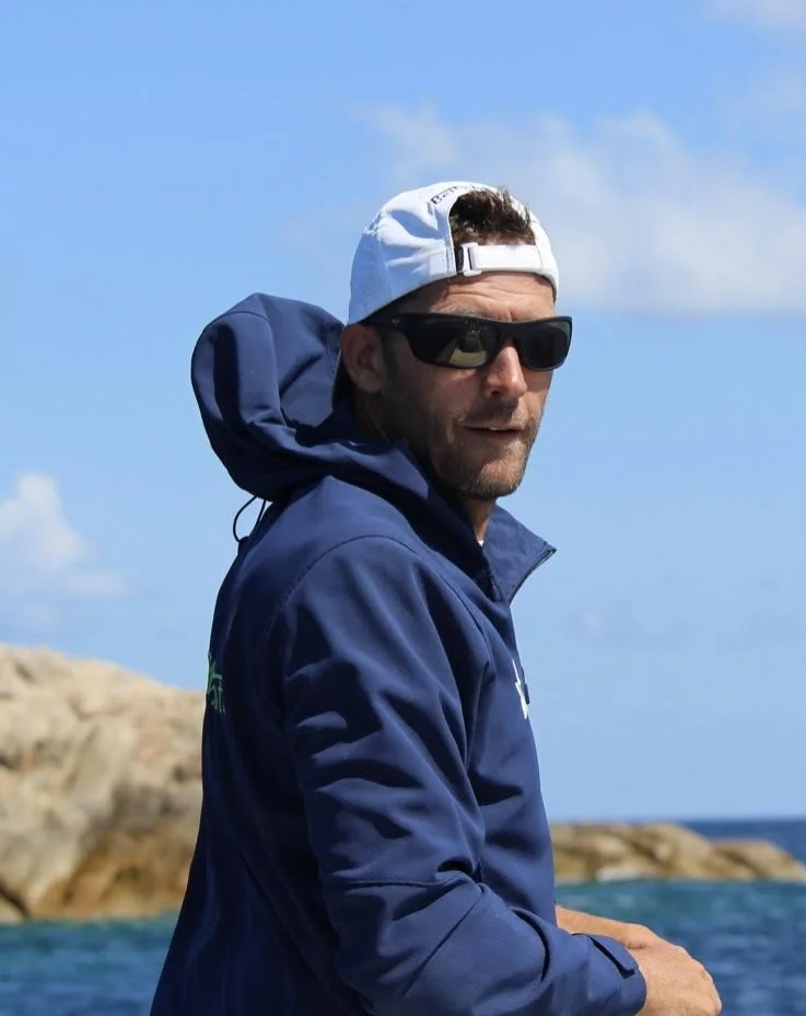 A man wearing a white baseball cap backwards, black sunglasses, and a blue jacket outdoors near water and rocks.