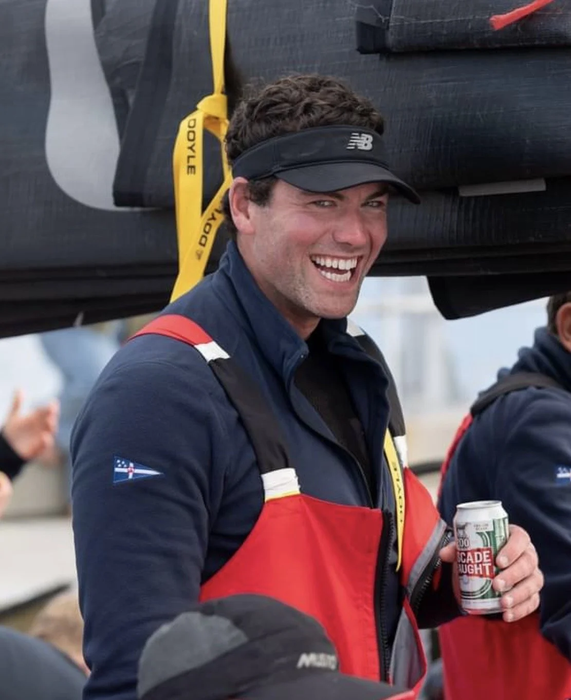 A man smiling while holding a can of beer, wearing a black sports visor, a dark blue jacket with red and white accents, and a red life vest. He has a black backpack and is surrounded by others at an outdoor event.