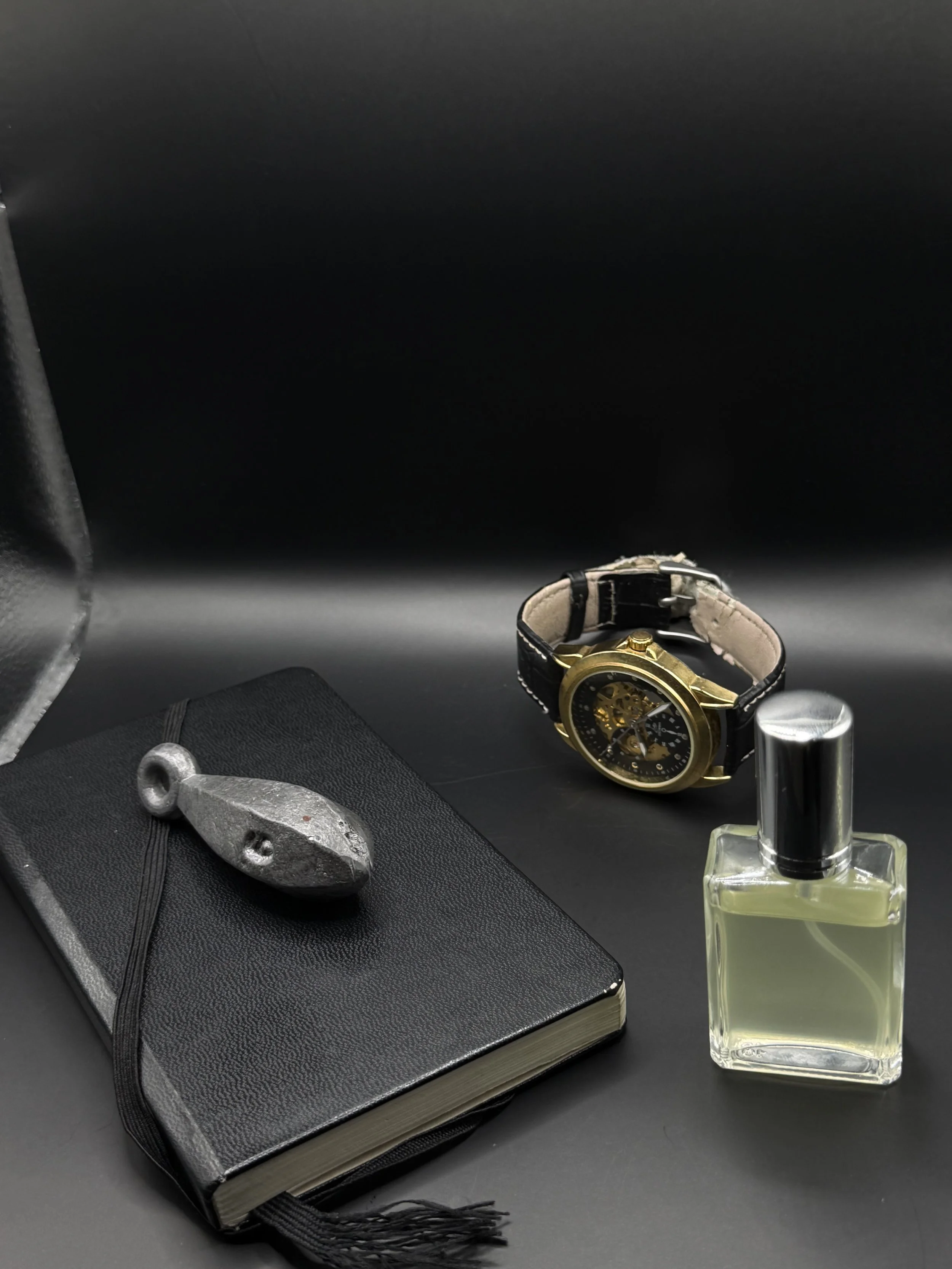 Black notebook with a pen on top, a small bottle of perfume, a wristwatch with a black strap and gold case