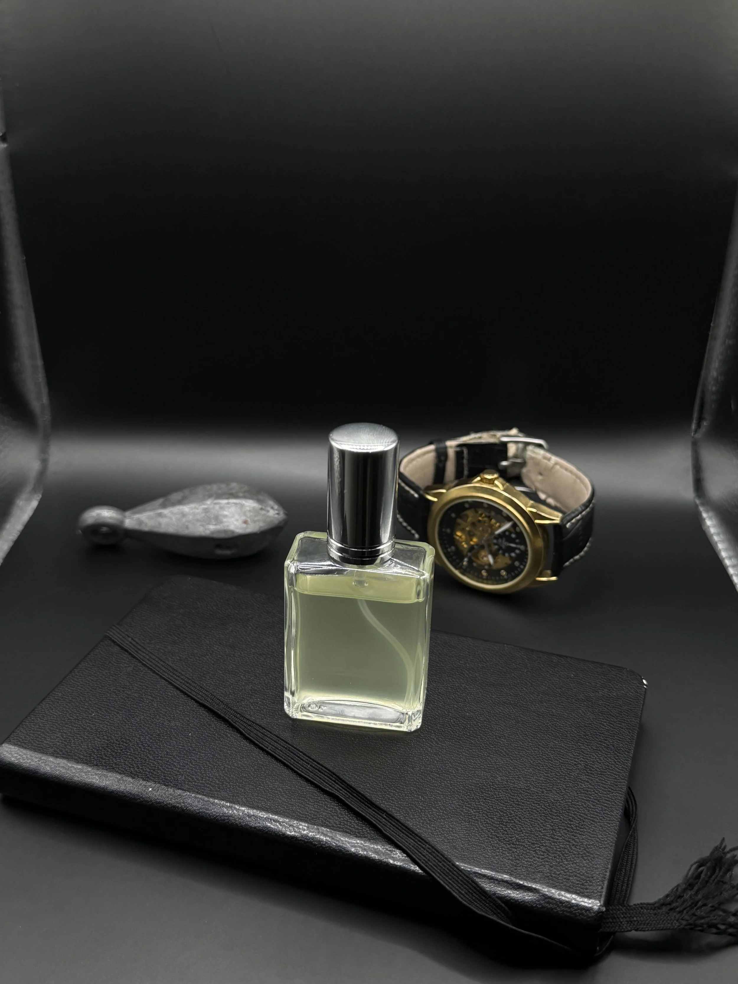 A black notebook, perfume bottle, gold and black wristwatch, and a gray rock are displayed on a black surface.