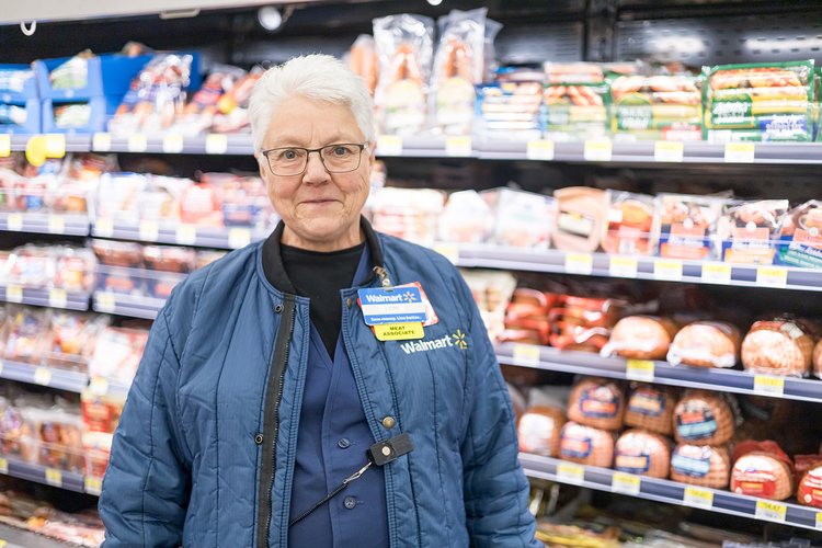 Walmart employee standing in meat aisle with packaged meats on shelves behind her.