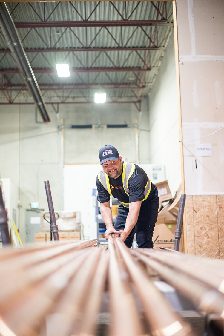A worker in a warehouse wearing a black and yellow safety vest and a black cap, smiling as he handles metal pipes surrounded by boxes and construction materials.