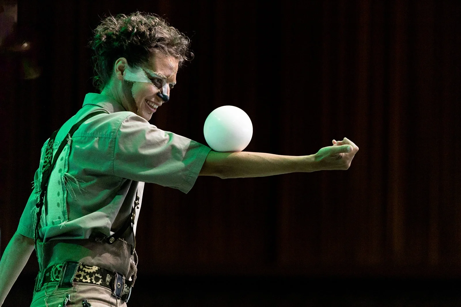 A performer with face paint resembling a cat, wearing a khaki shirt and patterned belt, balancing a glowing ball on their forearm in a performance setting with dark background.
