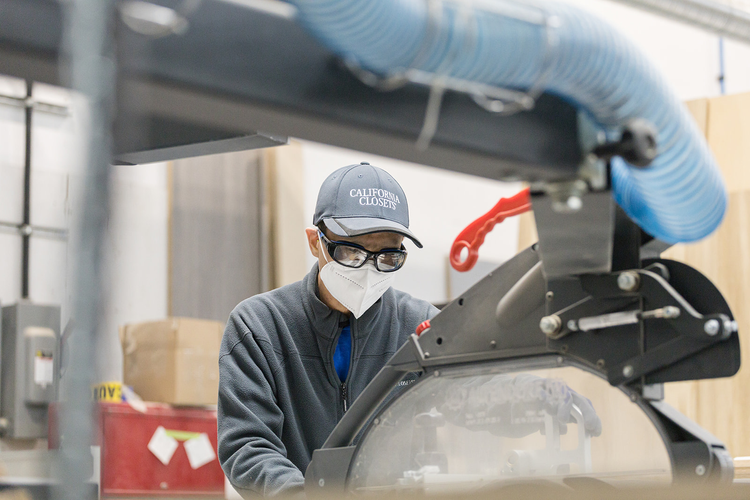 A worker wearing a face mask, safety glasses, a gray cap that says 'California Closets', and a gray jacket operating machinery in a warehouse or workshop.