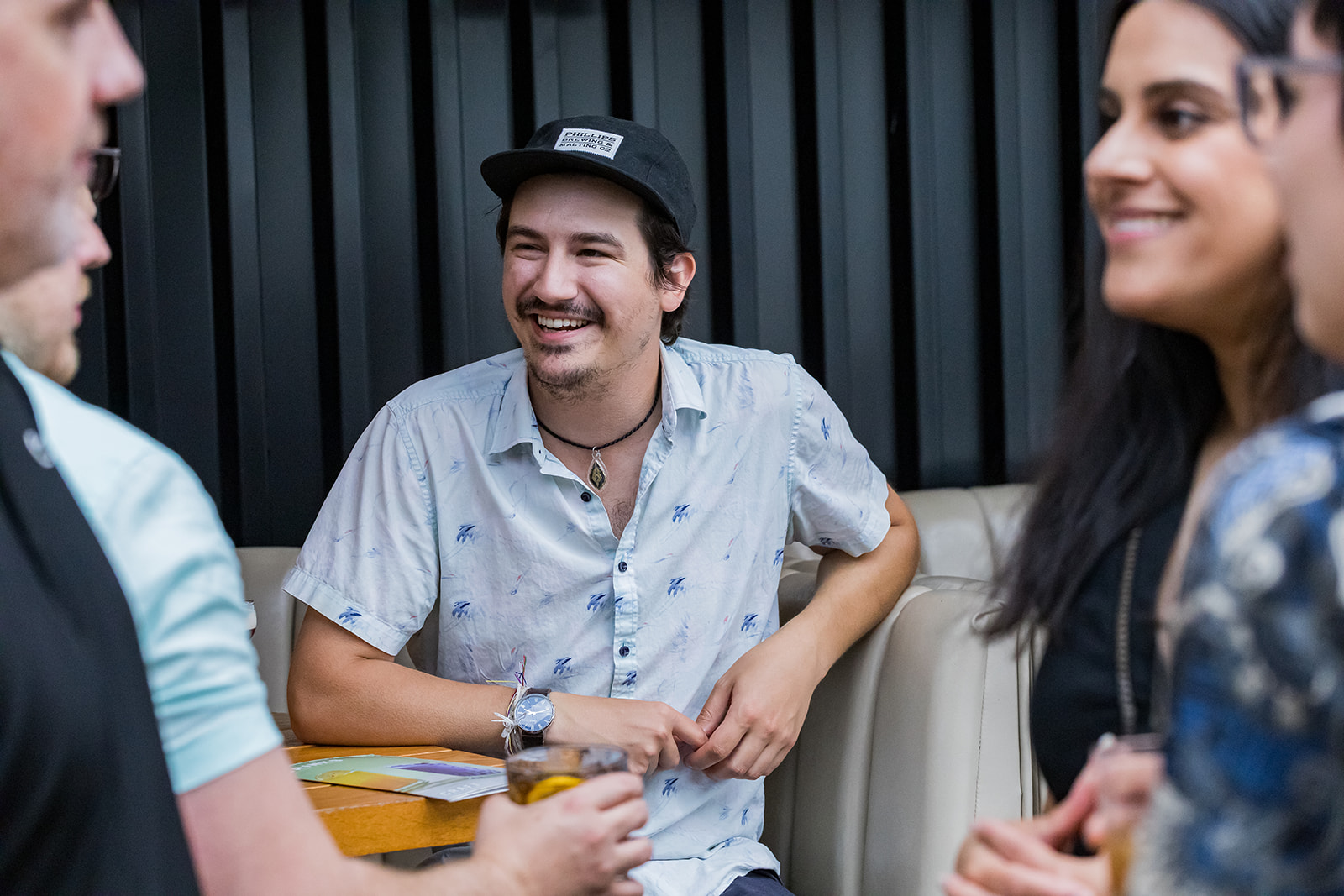 A group of young adults enjoying a social gathering, with one man smiling and talking, holding a drink, and others engaged in conversation.