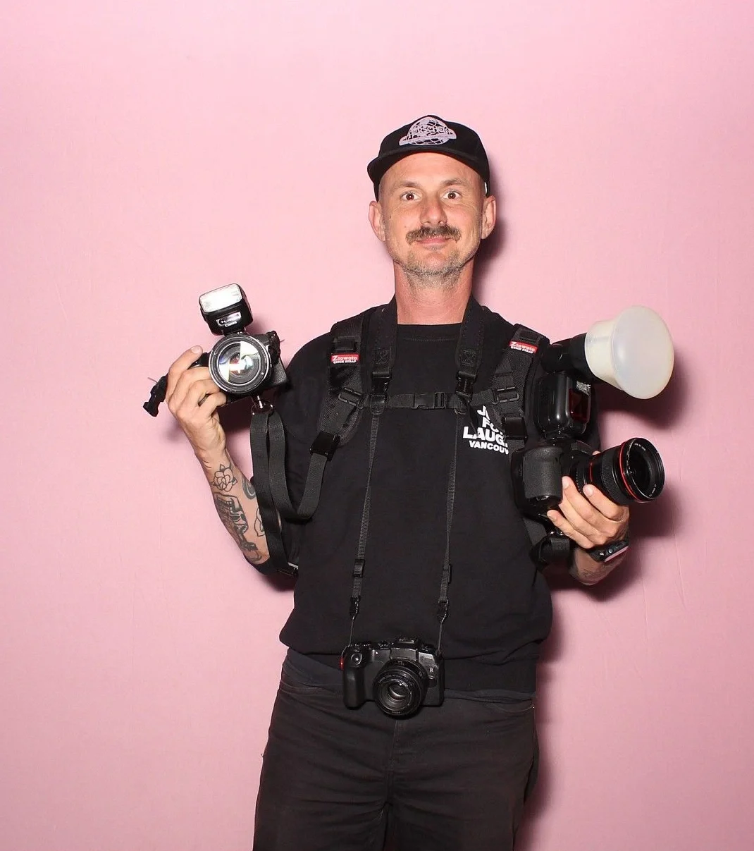 Ryan Walter Wagner portrait against pink background, holding professional camera equipment, wearing a black cap, black T-shirt with white text, and a camera hanging around his neck.