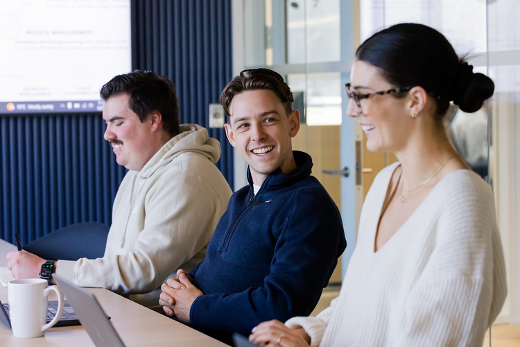 Three young adults sitting at a table in a modern office, engaged in conversation and smiling.