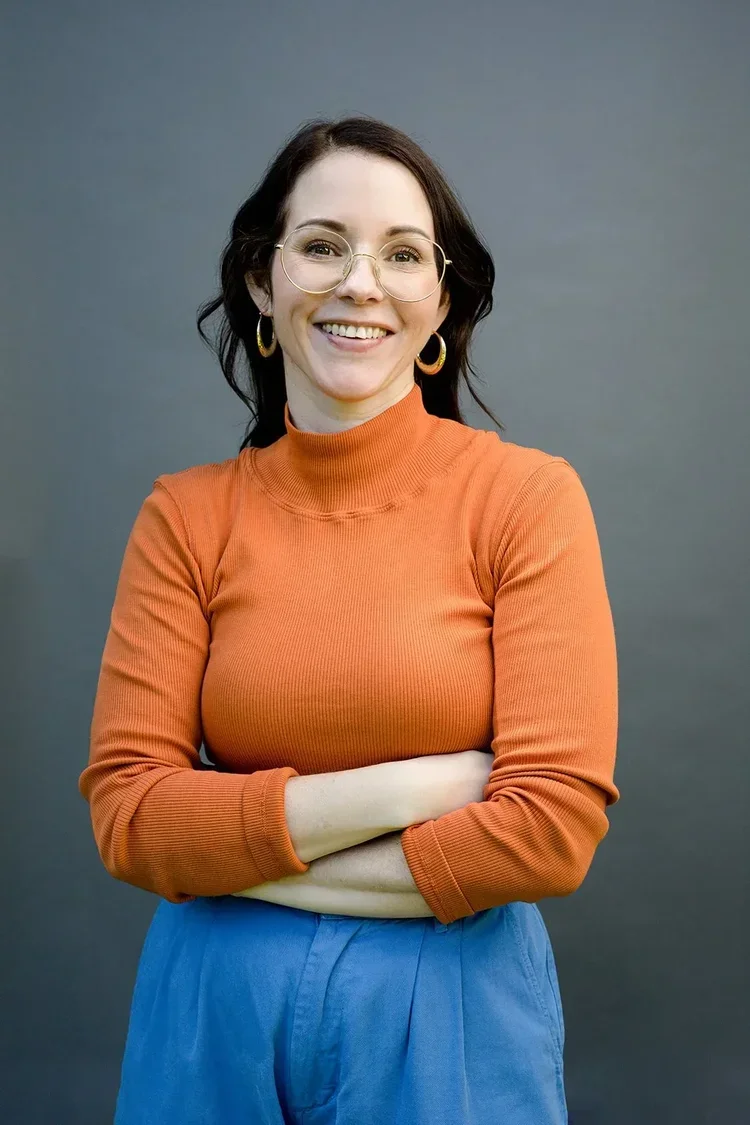 A woman with dark brown hair, glasses, and hoop earrings smiling with arms crossed, wearing an orange turtleneck sweater and blue pants against a plain gray background.
