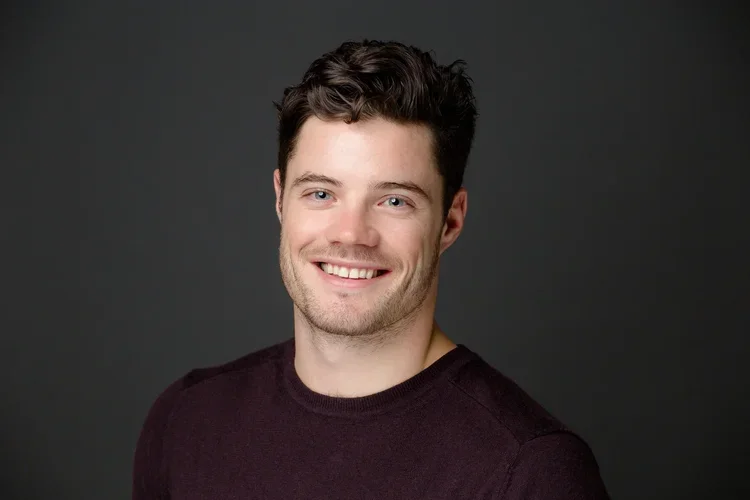 Portrait of a young man with dark hair and blue eyes, smiling, wearing a dark shirt, against a gray background.