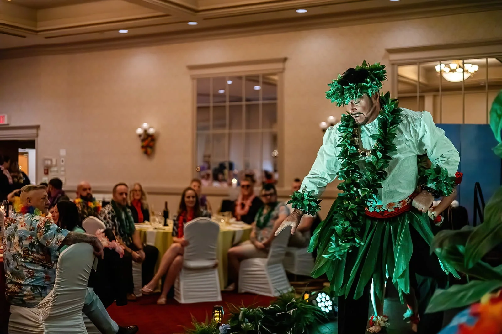 A man dressed in a green Hawaiian-themed costume with a lei and hat, performing on stage at a tropical-themed party with guests sitting at tables in the background.