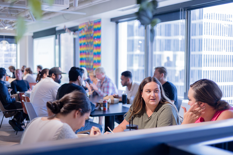 People sitting and talking at tables in a bright, modern restaurant or cafeteria with large windows and colorful artwork on the wall.