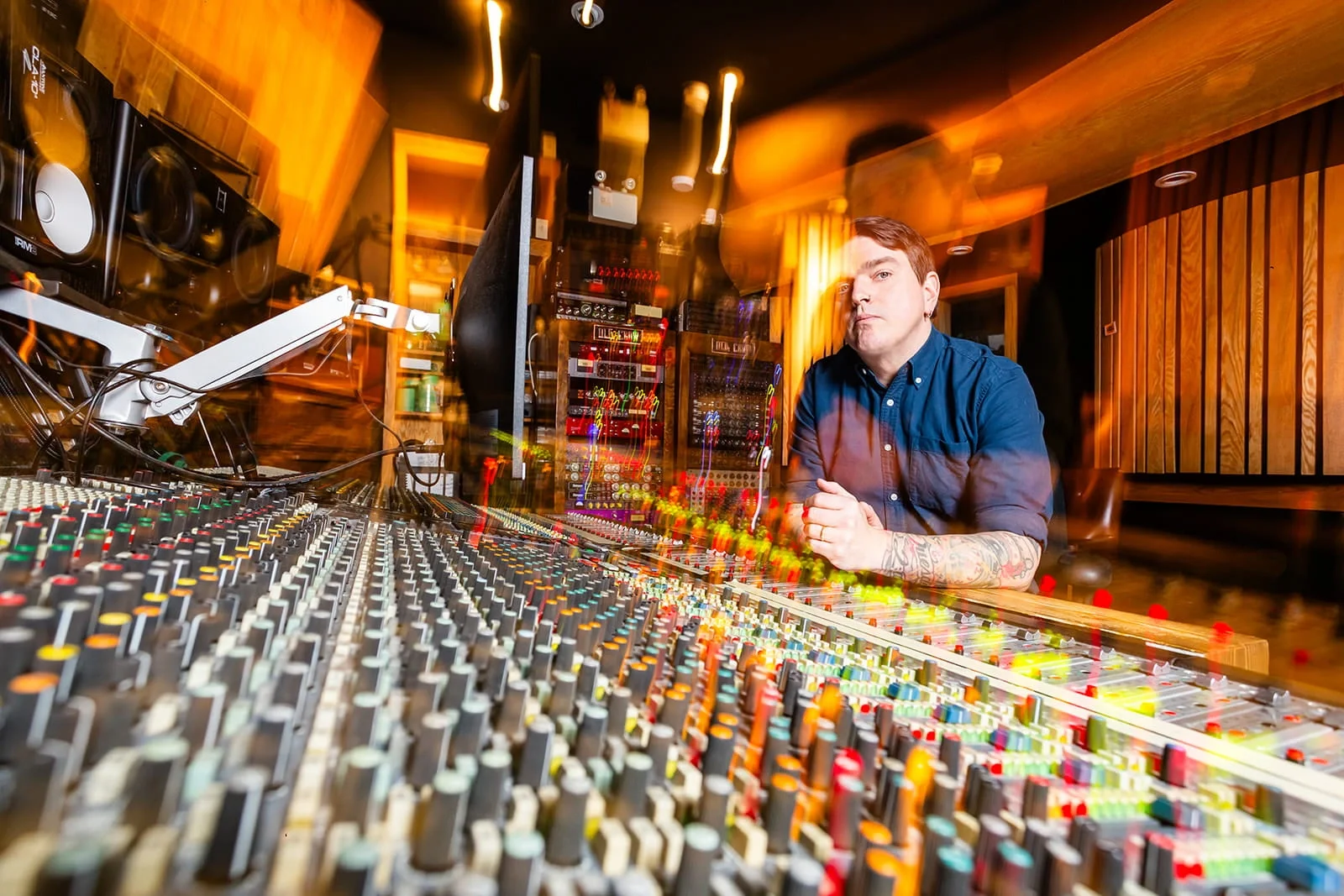 A man with tattoos on his forearm is sitting at a large audio mixing console in a recording studio, with sound equipment and wooden wall panels visible in the background.